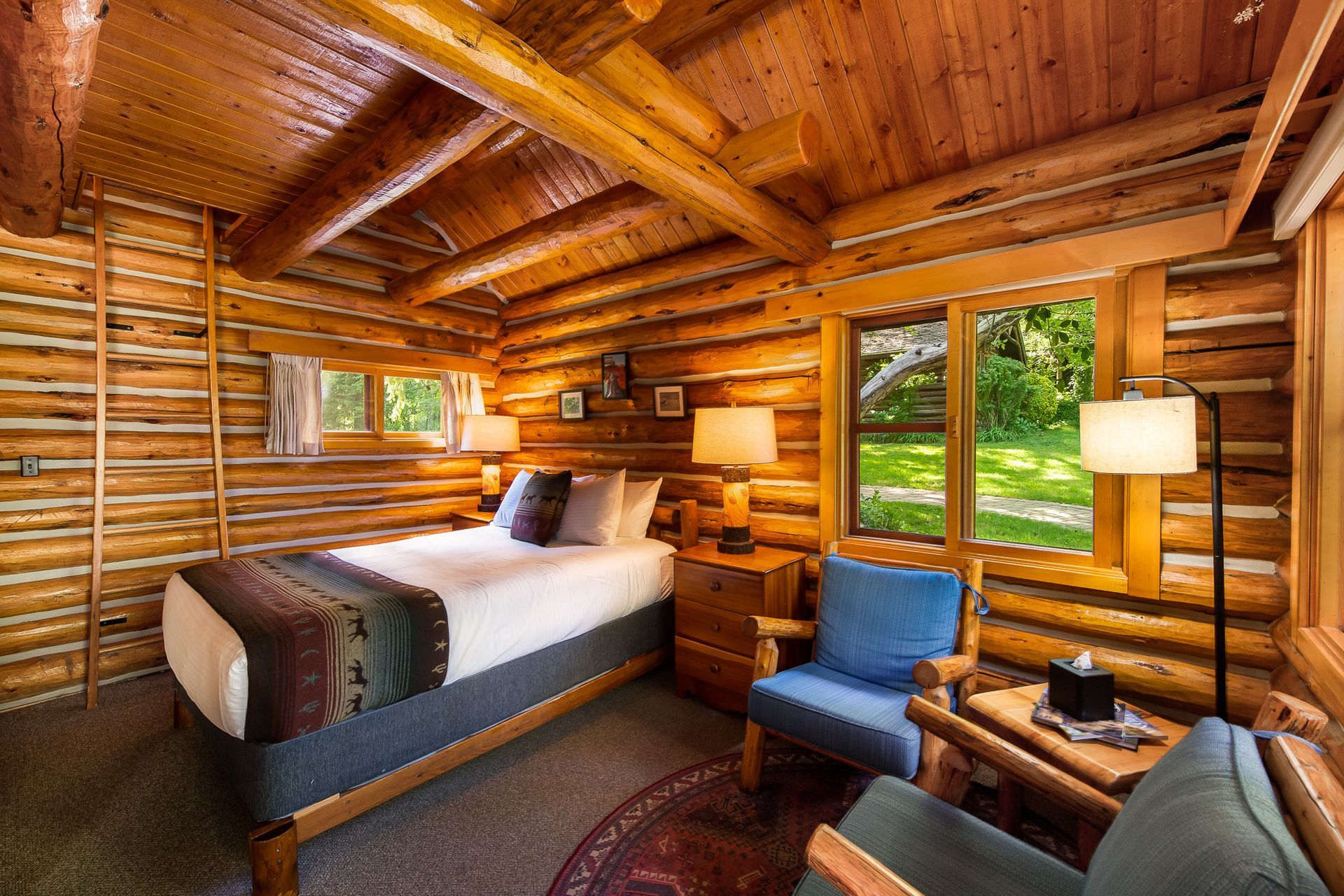 Rustic log cabin bedroom with wooden walls, bed, blue chair, lamp, and forest view through window