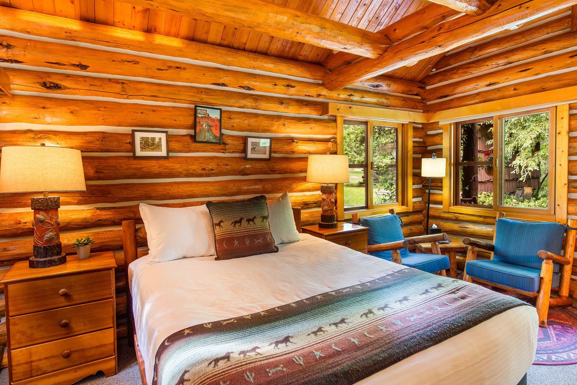 Cozy log cabin bedroom with a bed, bedside lamps, blue chairs, and large windows overlooking trees