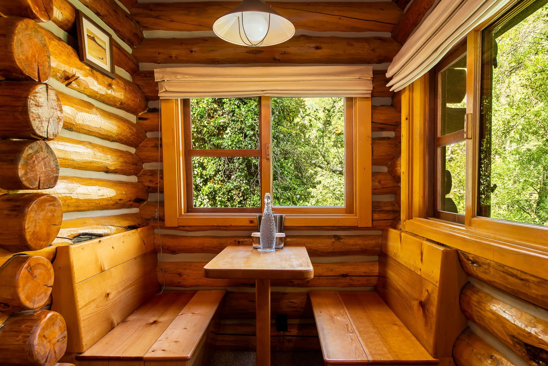 Cozy wooden cabin dining nook with log walls, a table, benches, and sunlit windows overlooking greenery