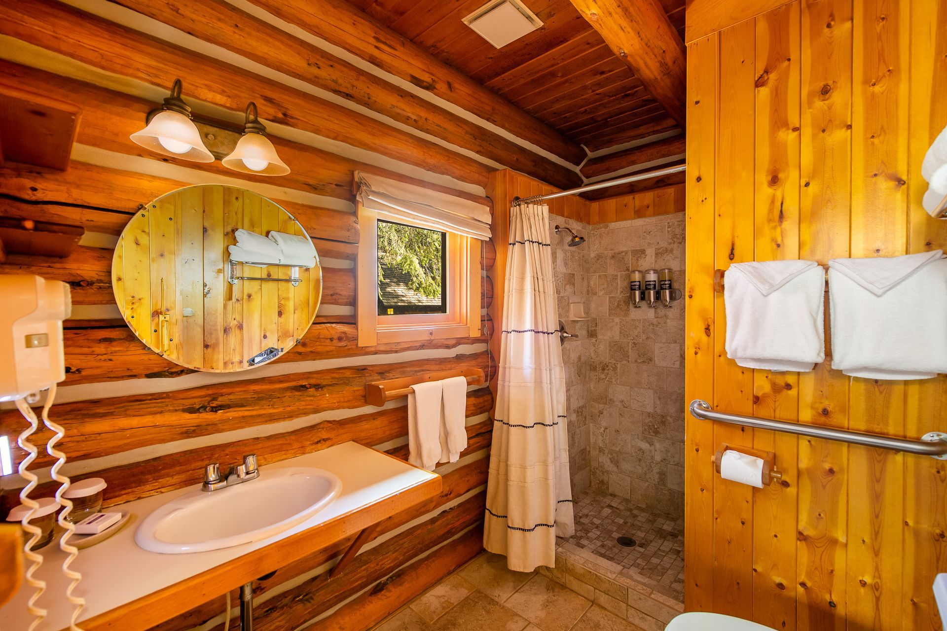 Rustic log cabin bathroom with wood-paneled walls, round mirror, sink, shower curtain, and towels.