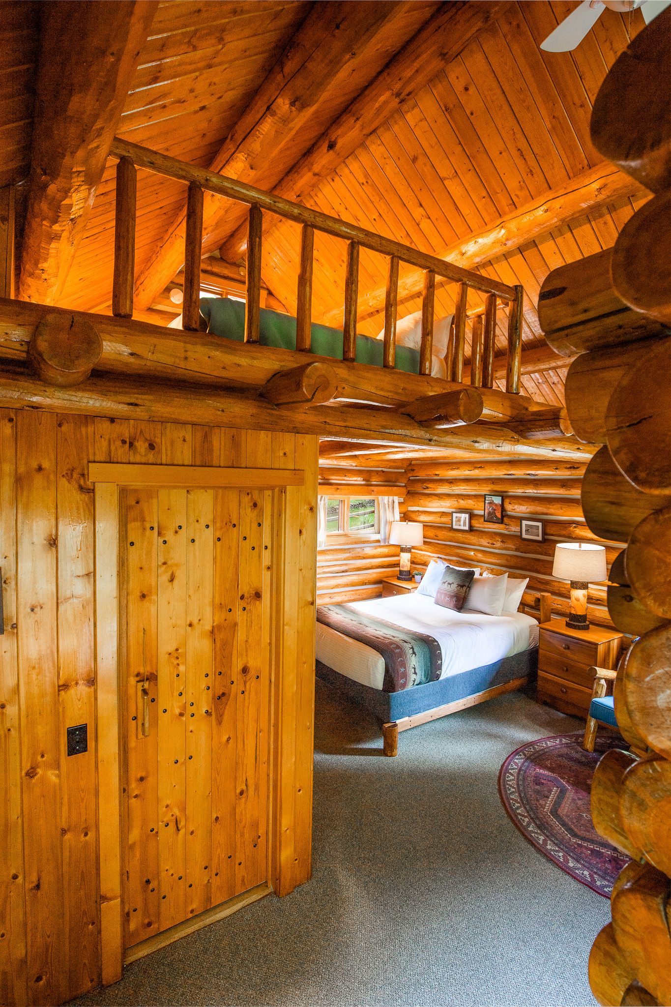 Rustic log cabin bedroom with wooden walls, loft, and a blue-covered bed by the window.