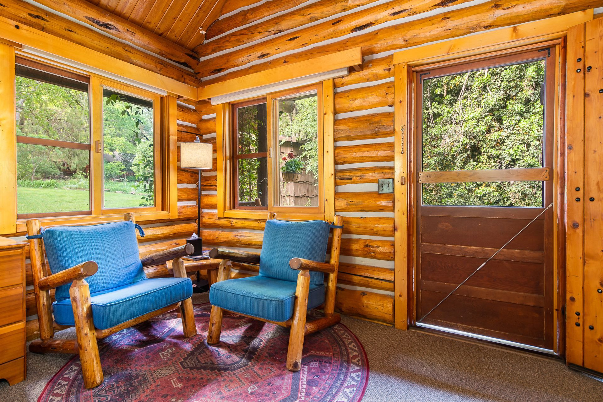 Log cabin interior with blue chairs, wood walls, and large windows looking out to greenery