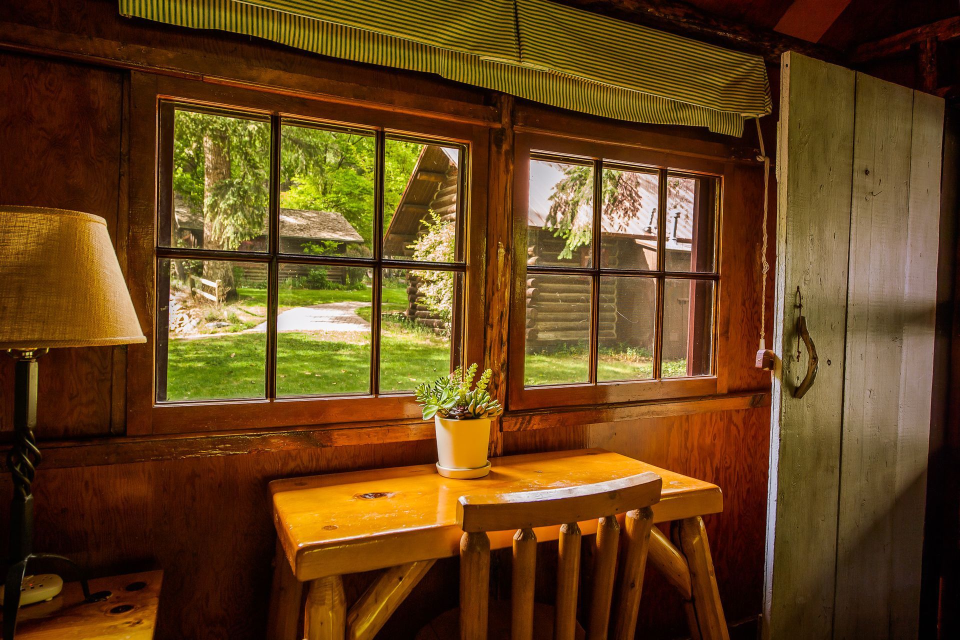 Cozy wooden room with a sunlit window, table, chair, and lamp overlooking greenery