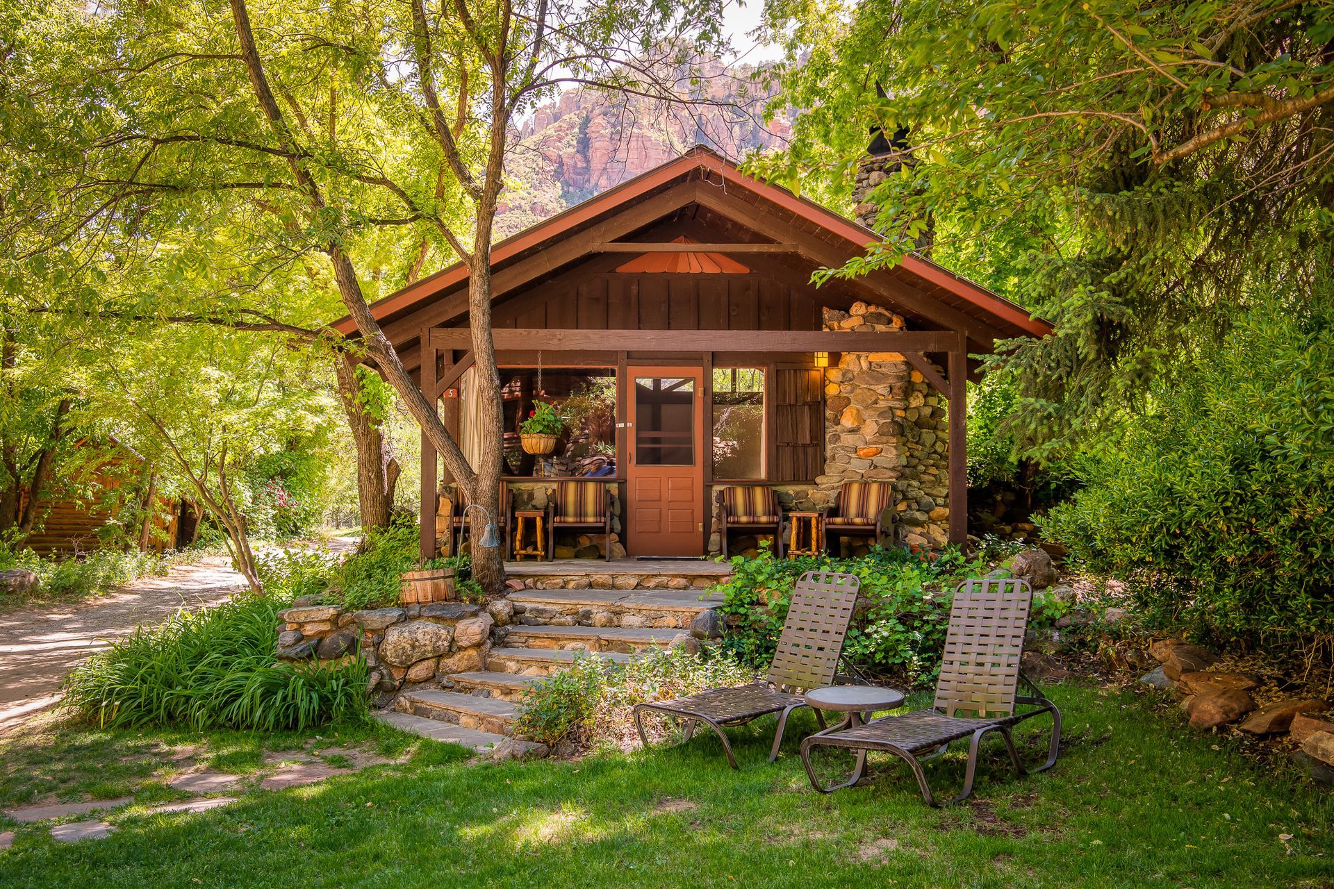 Cozy wooden cabin with porch and stone steps in a lush garden, surrounded by trees and lawn.