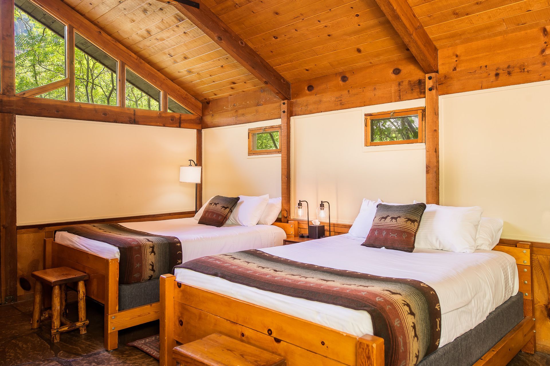 Cozy wooden cabin bedroom with two twin beds, warm lighting, and large windows overlooking greenery