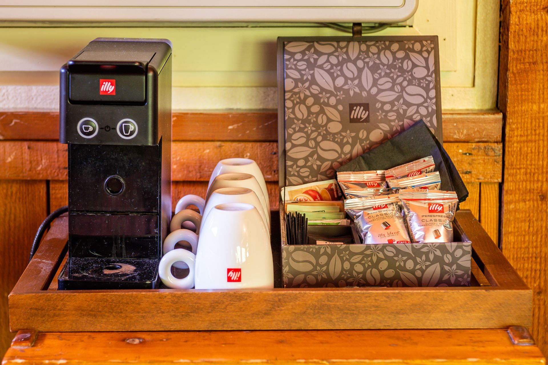 Coffee station with a black machine, cups, and assorted tea and coffee packets in a wooden tray.
