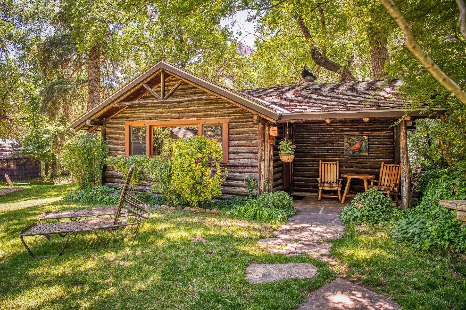 Rustic log cabin with a porch and rock path, surrounded by green trees and sunlight