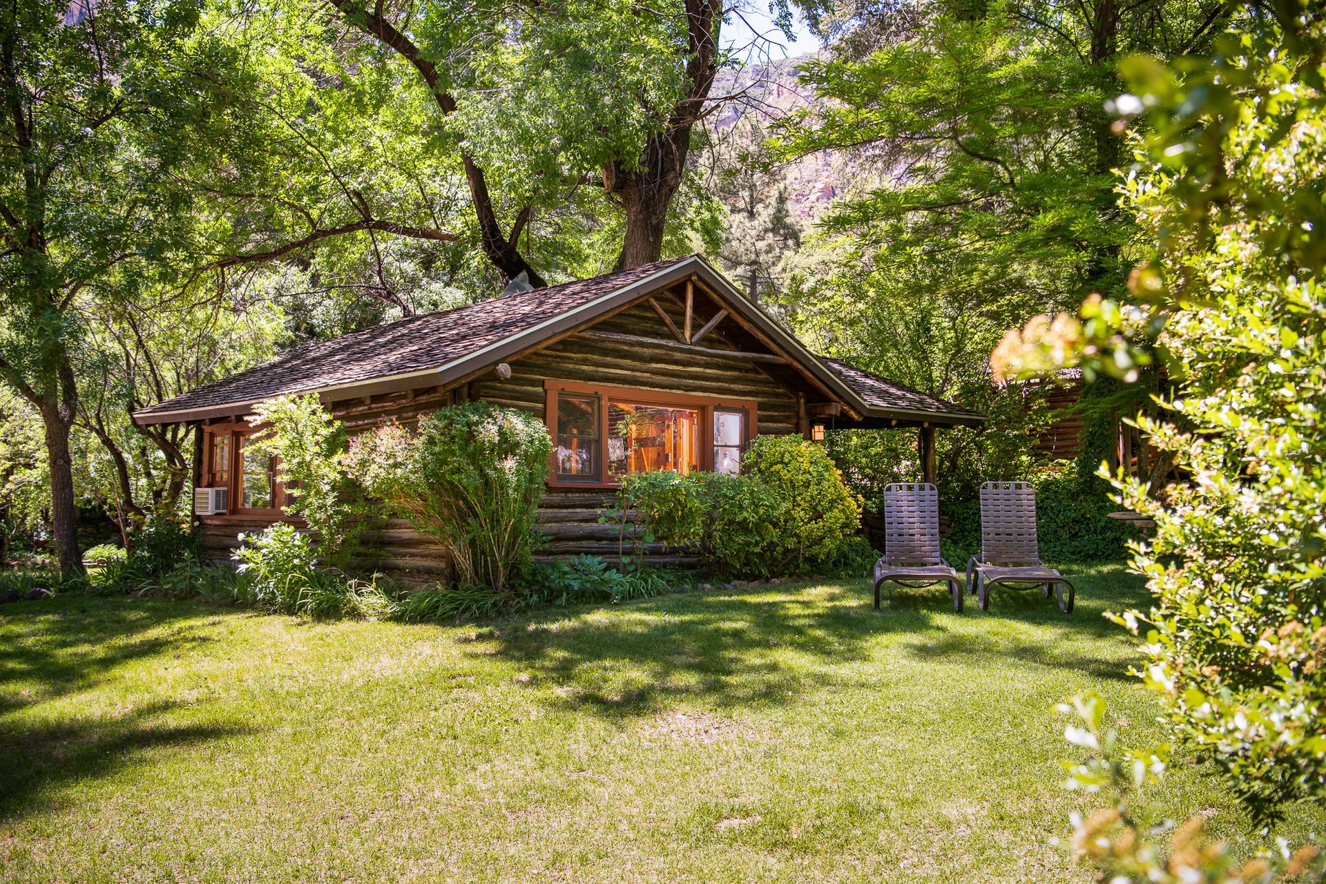 Rustic cabin in a lush green garden, surrounded by trees, with two chairs on the lawn.