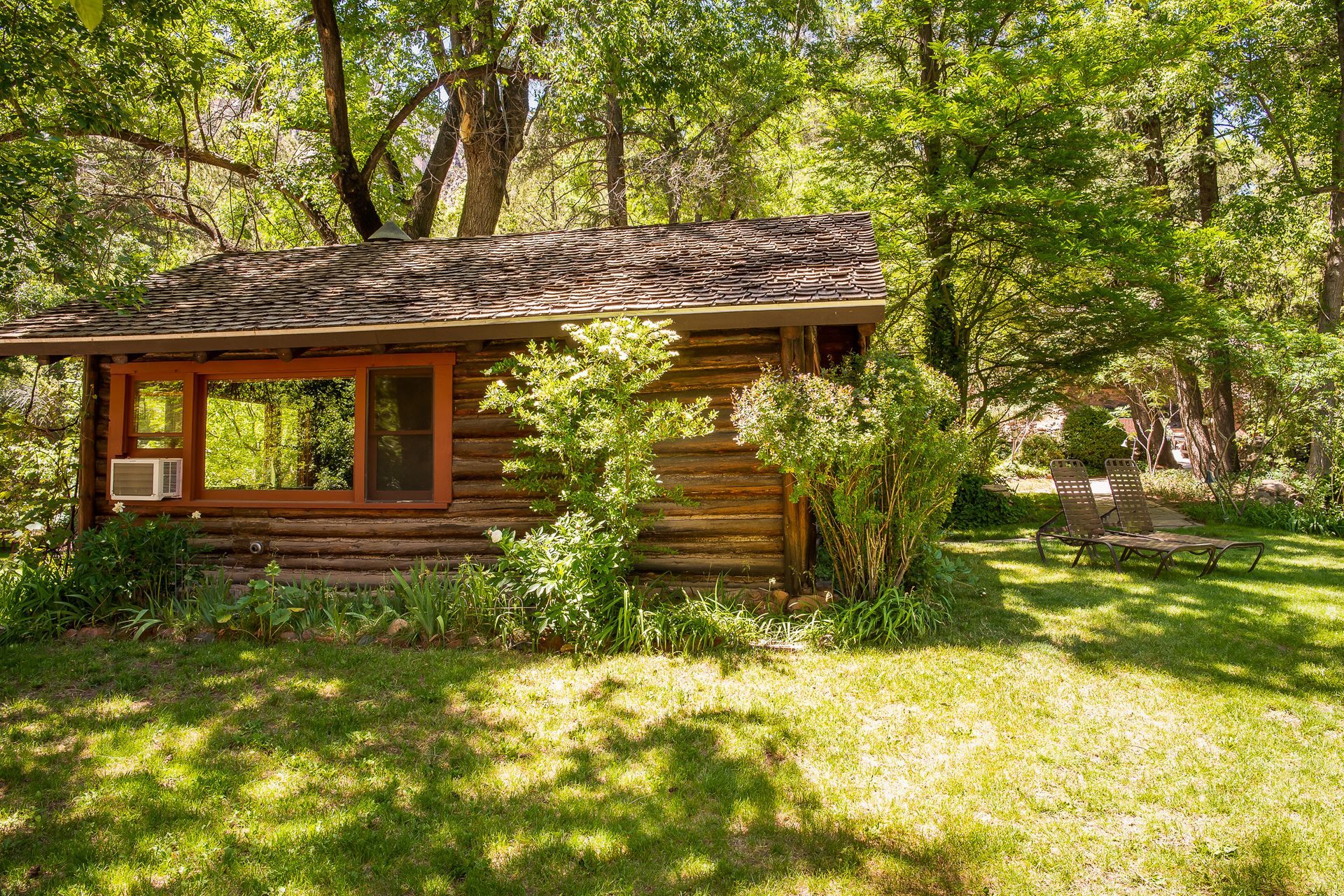 Small rustic log cabin in a sunlit wooded yard with green grass and trees