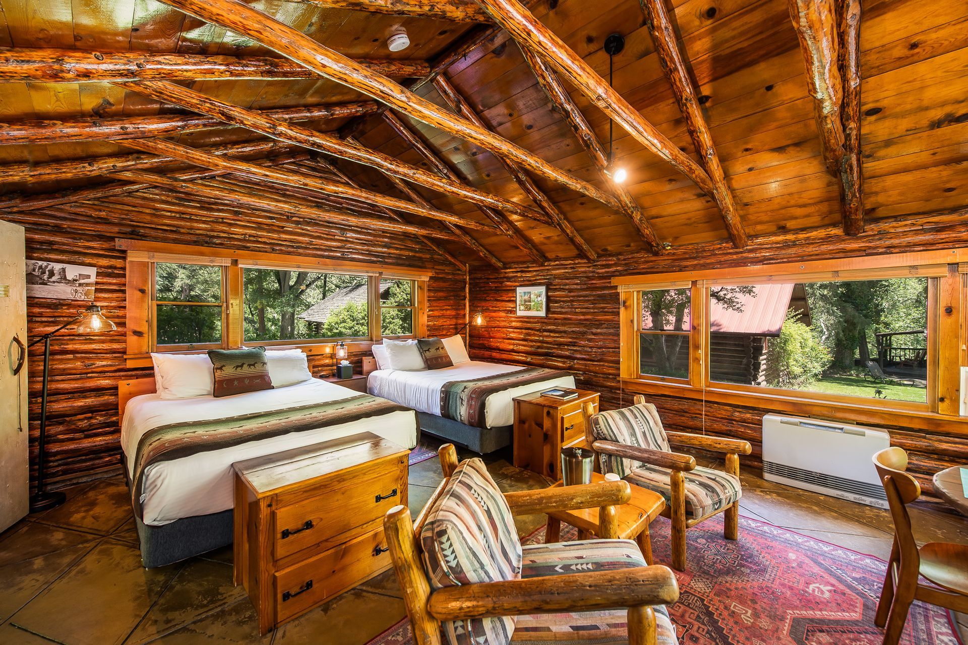 Rustic log cabin bedroom with two beds, wood beams, and large windows overlooking trees