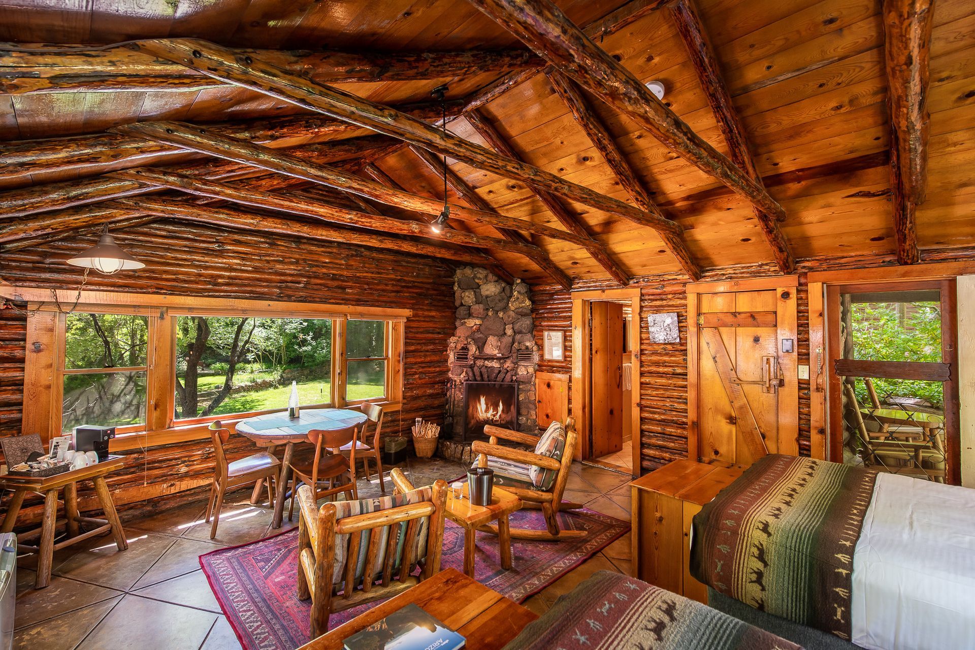 Rustic log cabin interior with wood beams, cozy seating, stone fireplace, and large windows overlooking trees