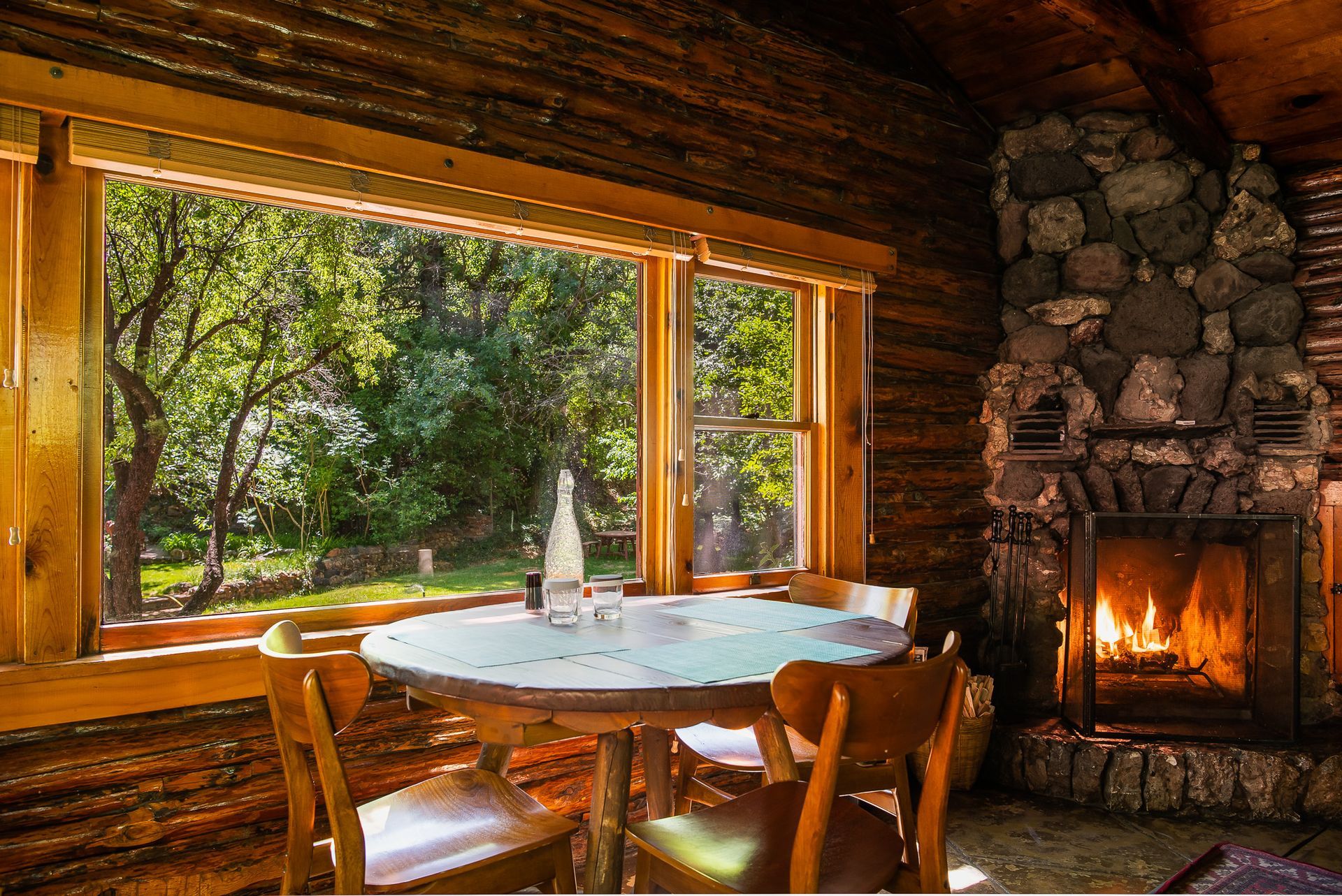 Rustic cabin dining nook with round table, wooden chairs, stone fireplace, and forest view through a large window