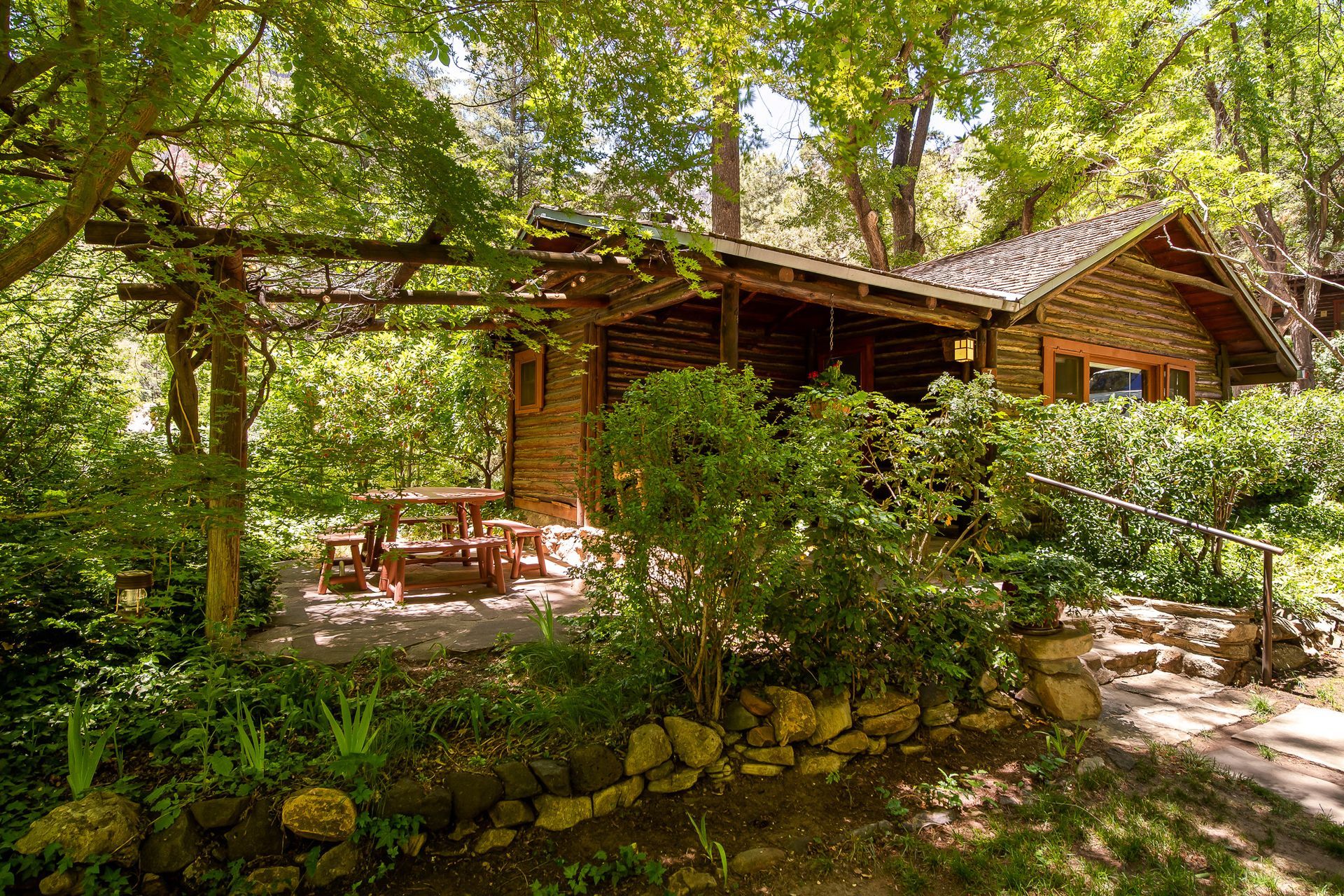 Wooden cabin in a lush forest with a stone path, shaded by tall trees and greenery