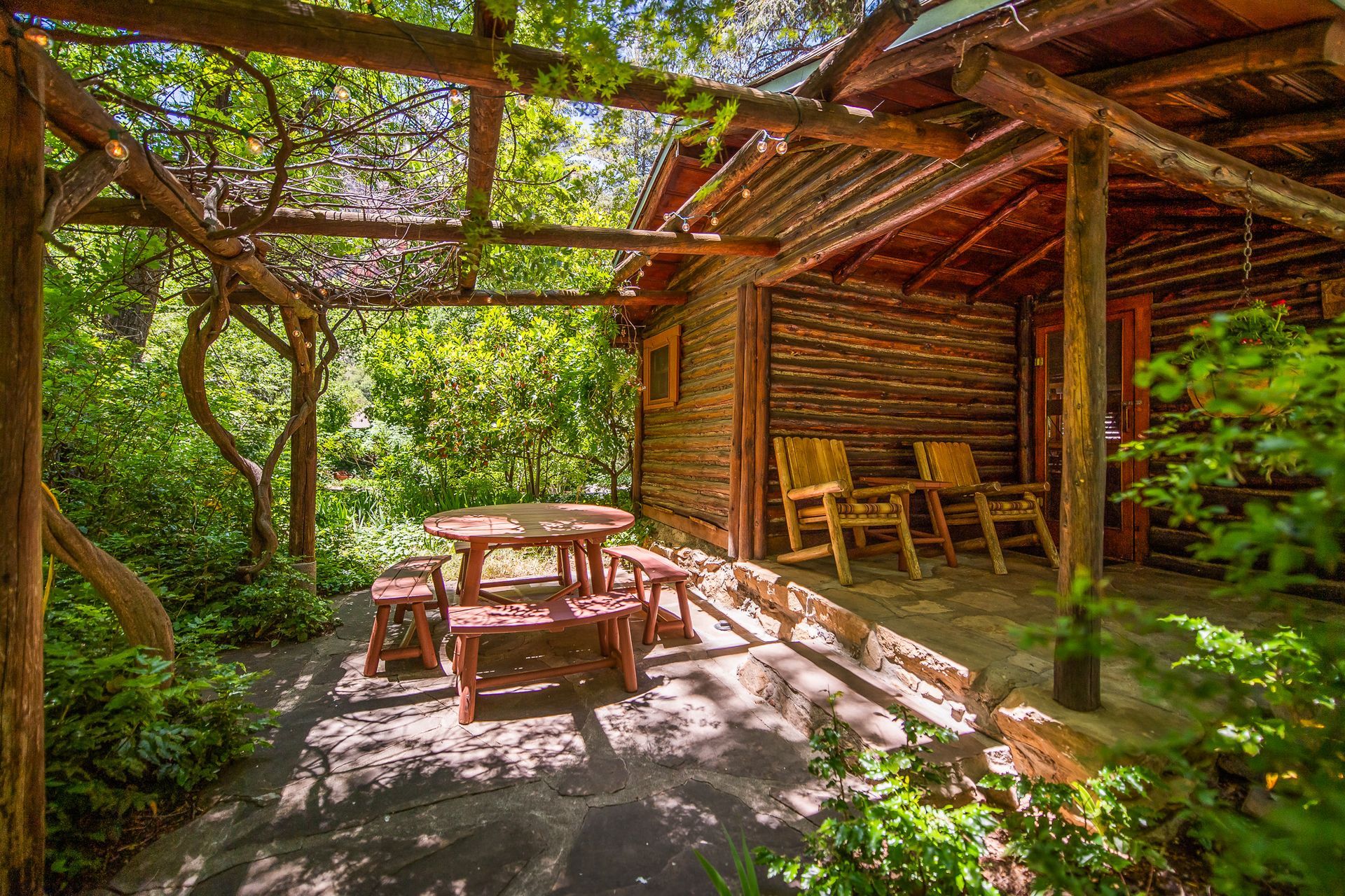Rustic outdoor patio with wooden table and chairs under a slatted roof, surrounded by lush greenery.
