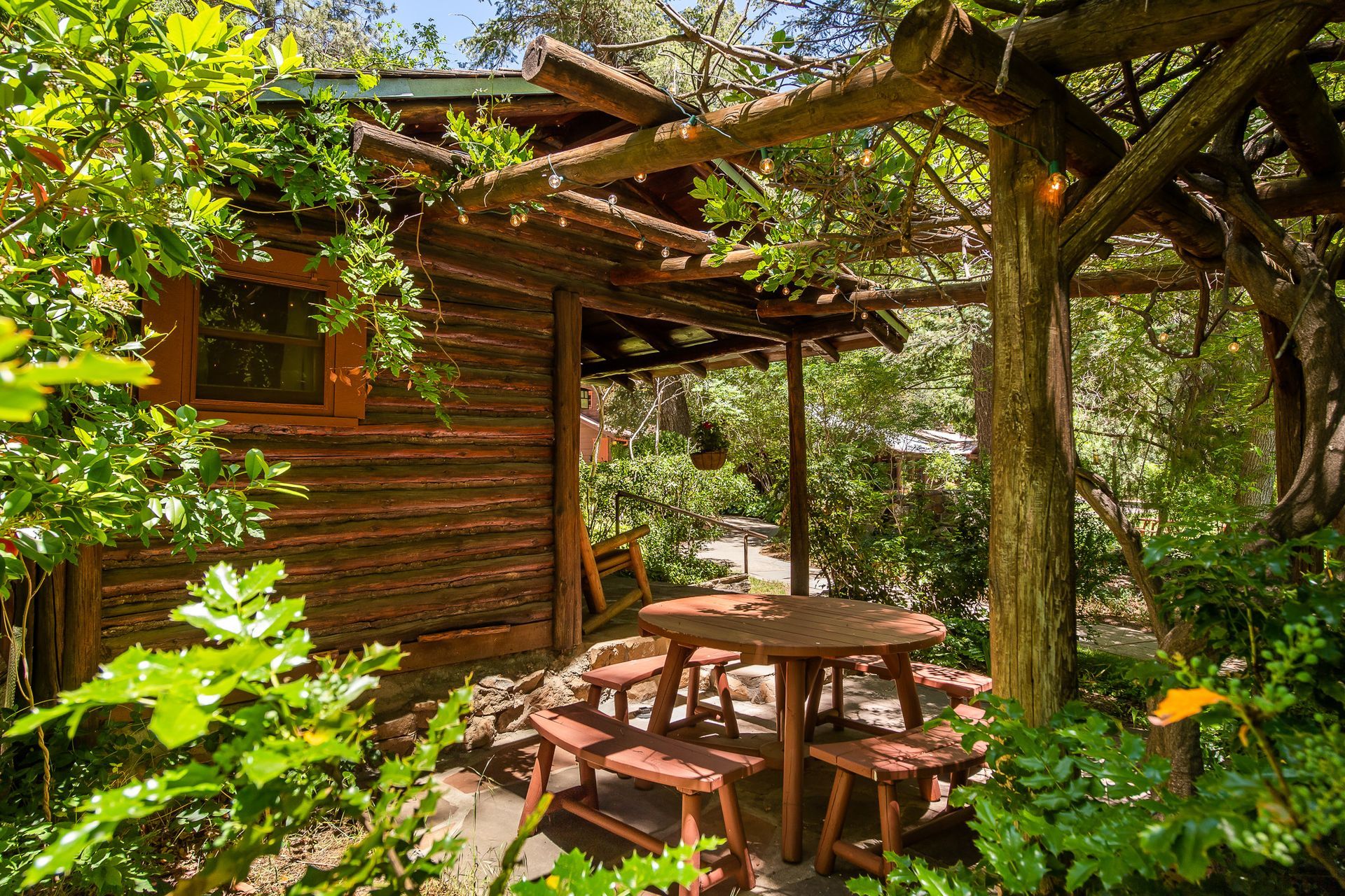 Wooden cabin patio with round table and benches, surrounded by lush green foliage and sunlight
