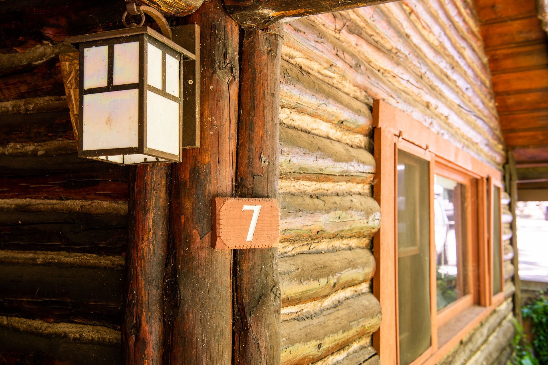 Rustic log cabin wall with lantern, red number 7 plaque, and a window.