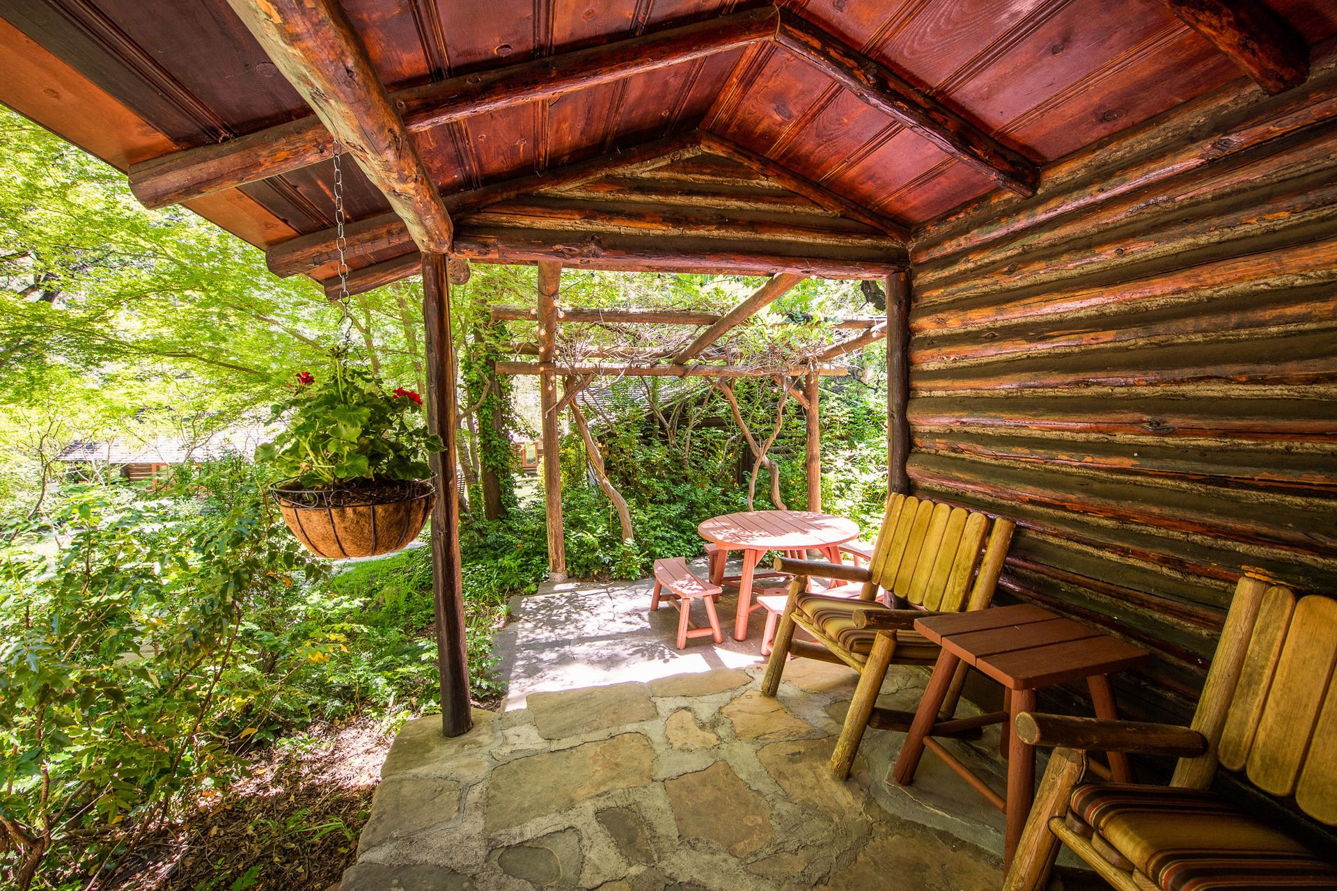 Rustic covered porch with wooden bench, table, hanging planter, and lush green garden view