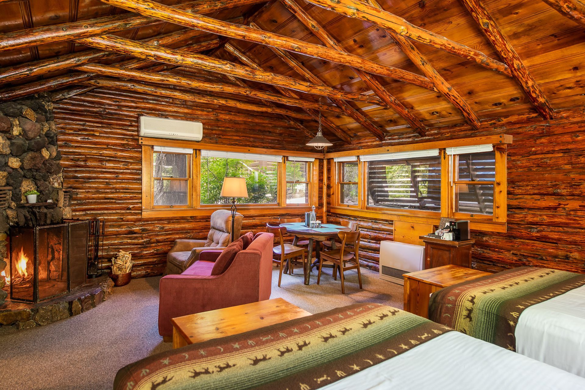 Rustic log cabin interior with a stone fireplace, seating area, and large windows overlooking trees