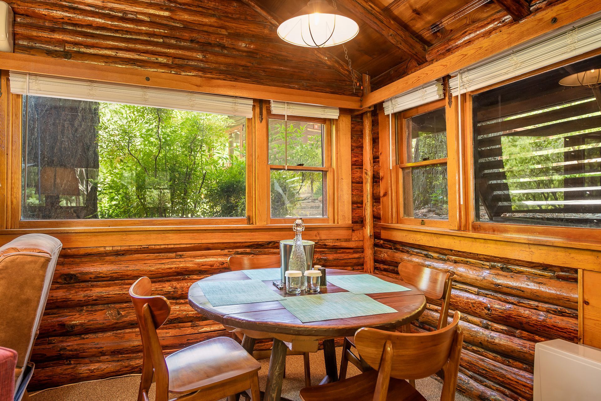 Rustic log cabin dining nook with round table, chairs, and large windows overlooking green trees
