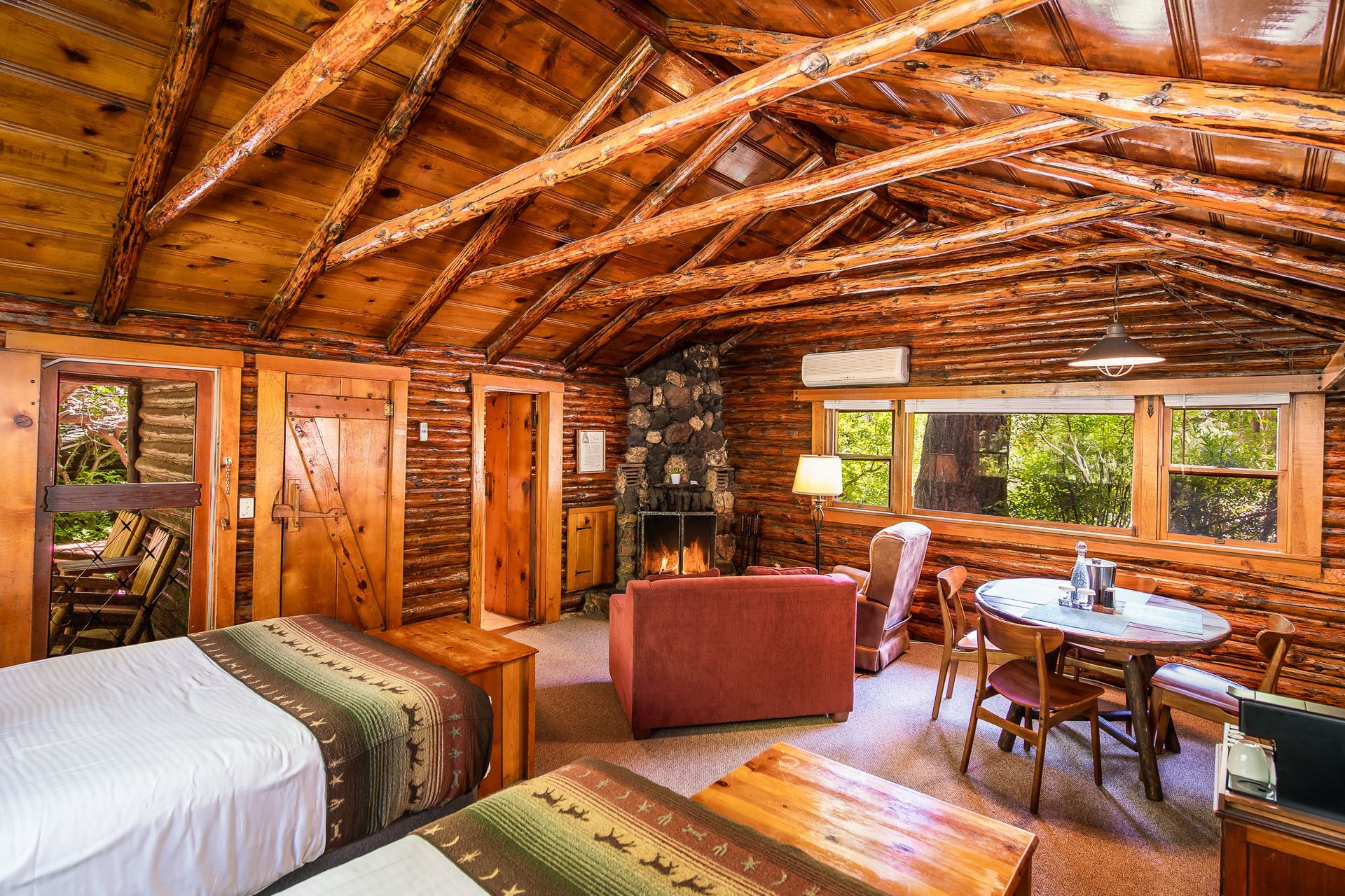 Rustic log cabin bedroom with exposed beams, bed, table, and large windows overlooking greenery