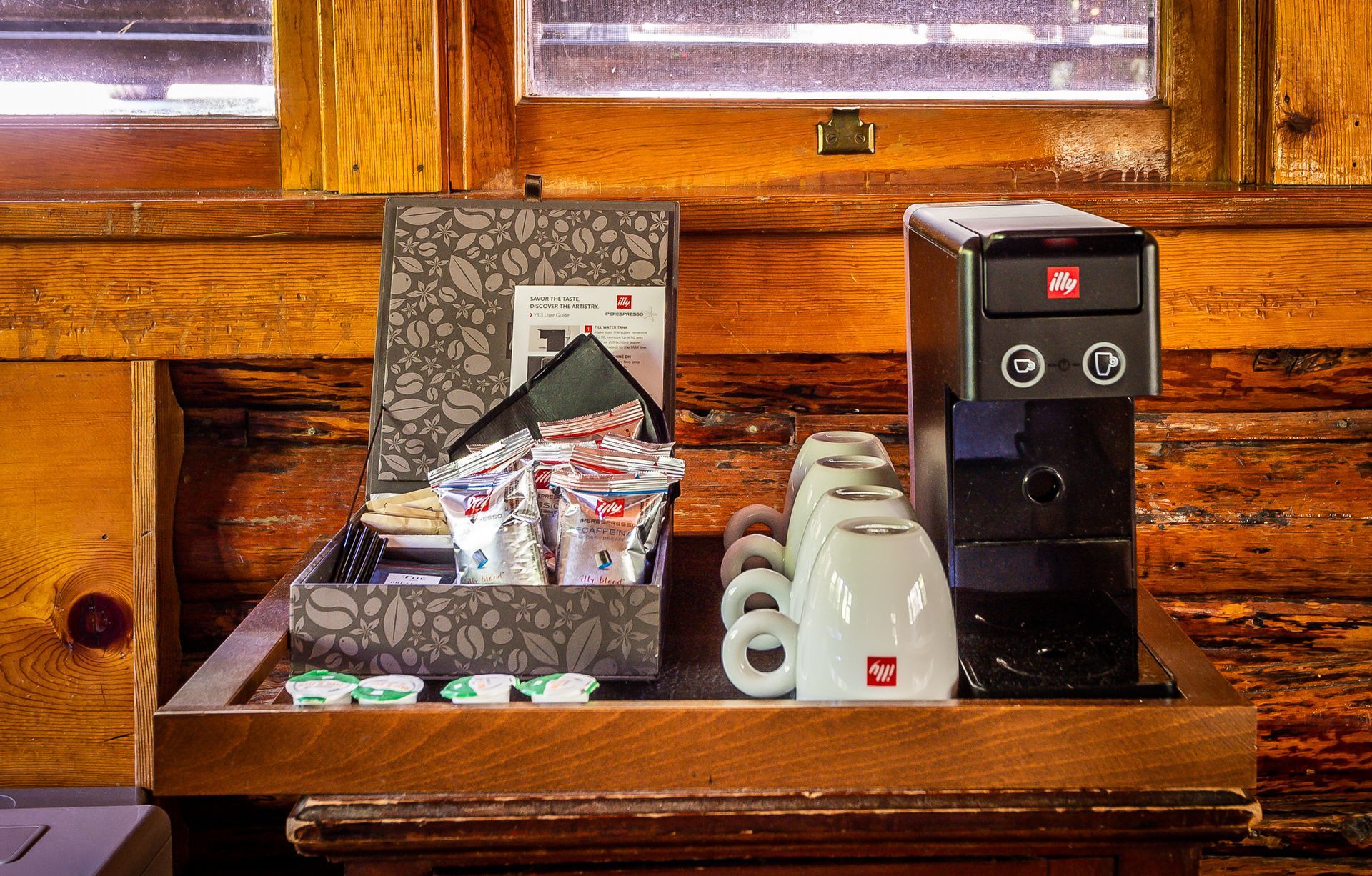 Coffee station with cups, napkins, and a beverage dispenser on a wooden shelf by a window
