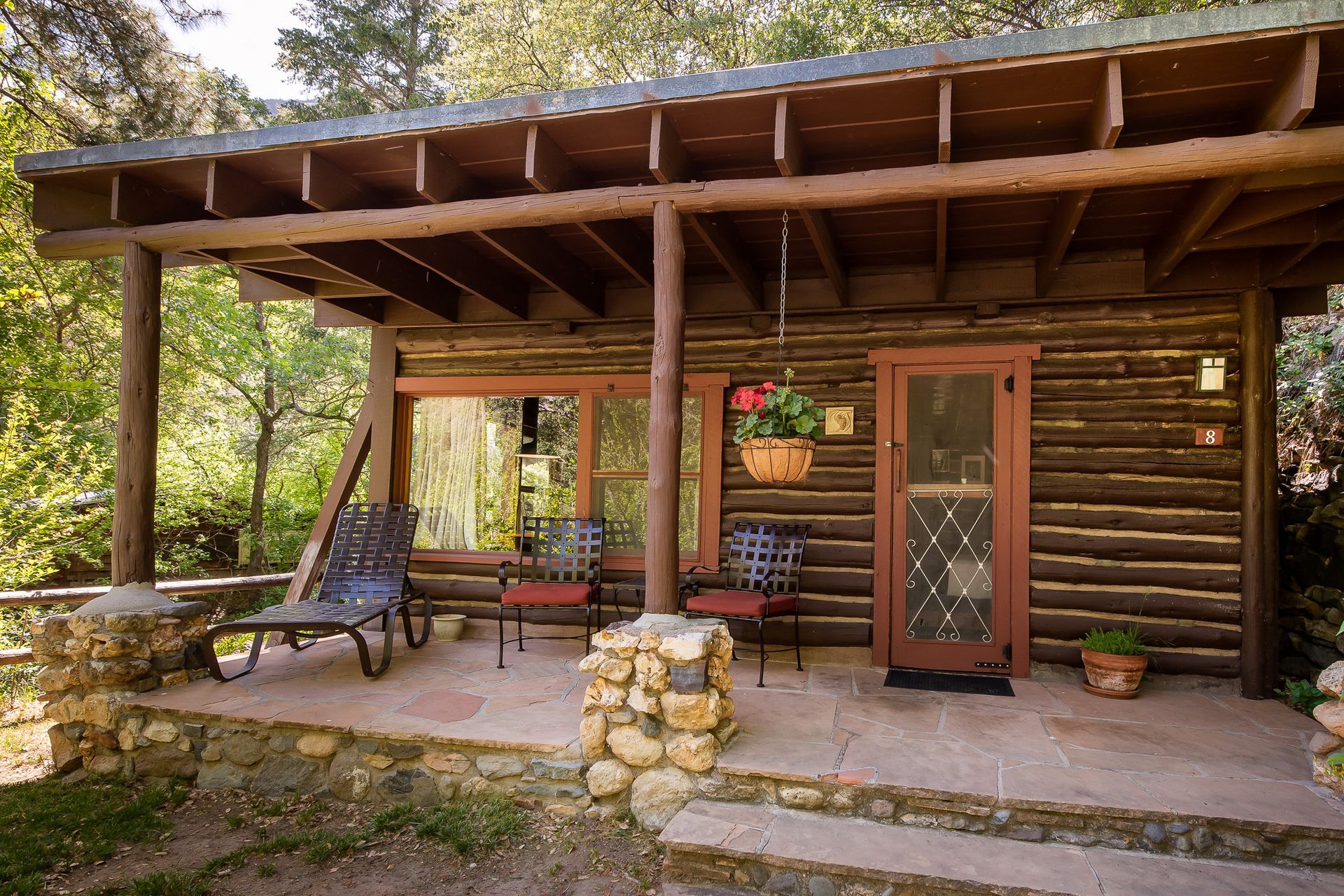 Rustic cabin porch with wooden chairs, hanging flowers, and a stone pile by the front door