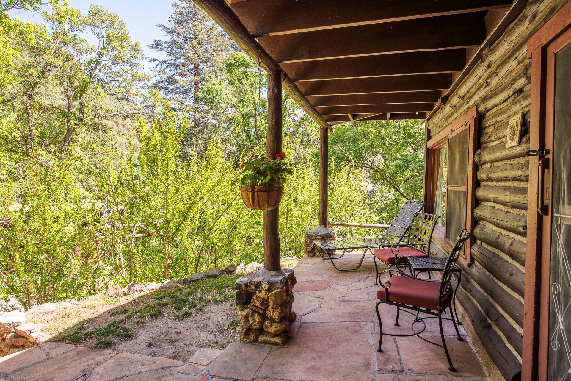 Rustic cabin porch with chairs, stone pillar, and greenery in warm sunlight