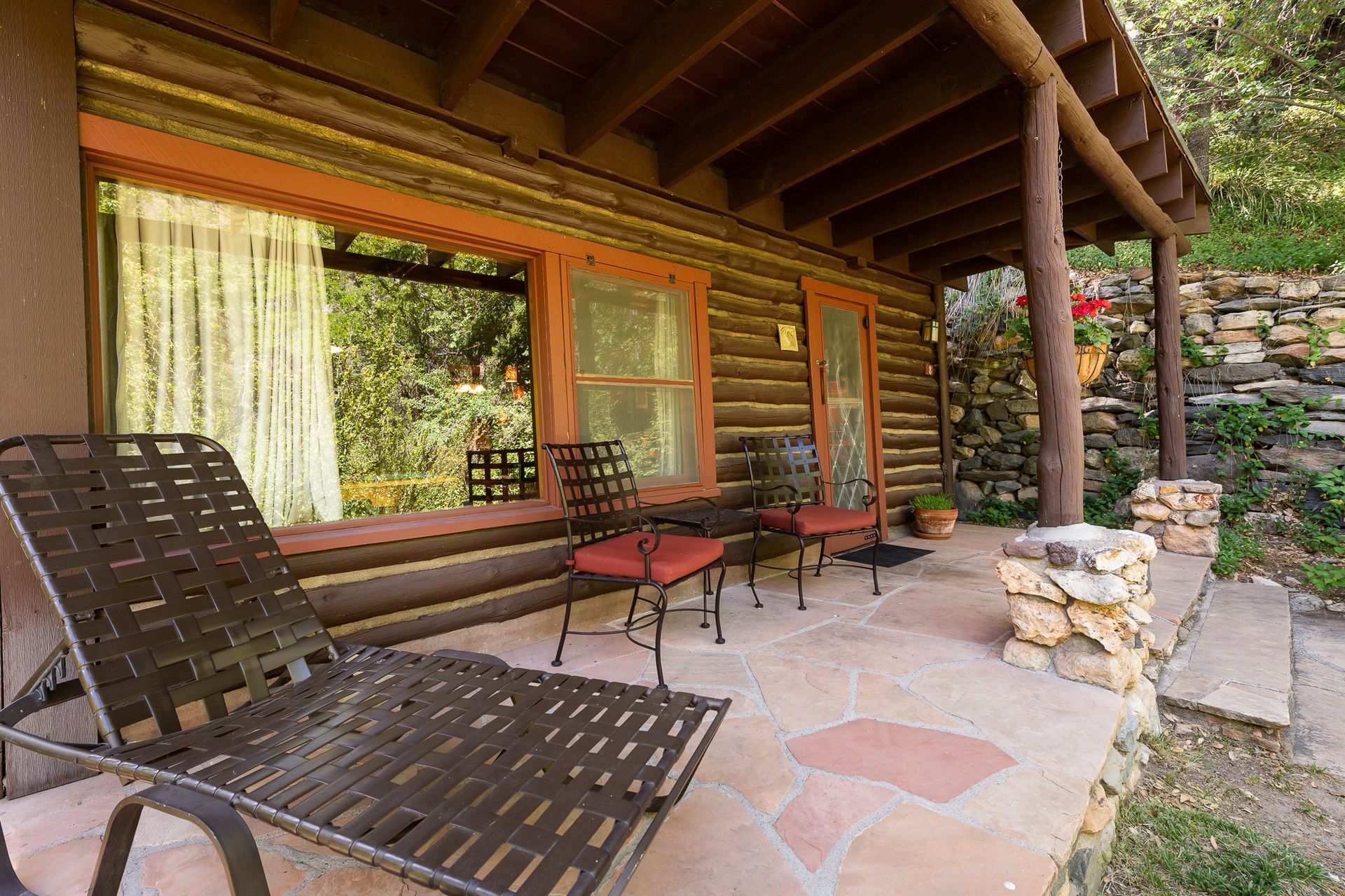 Log cabin porch with wooden lounge chairs, red table, stone fire pit, and large windows under a timber roof