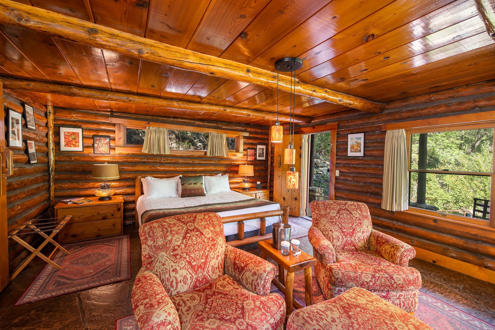 Cozy wooden cabin bedroom with bed, patterned armchairs, warm lighting, and a large window overlooking trees.