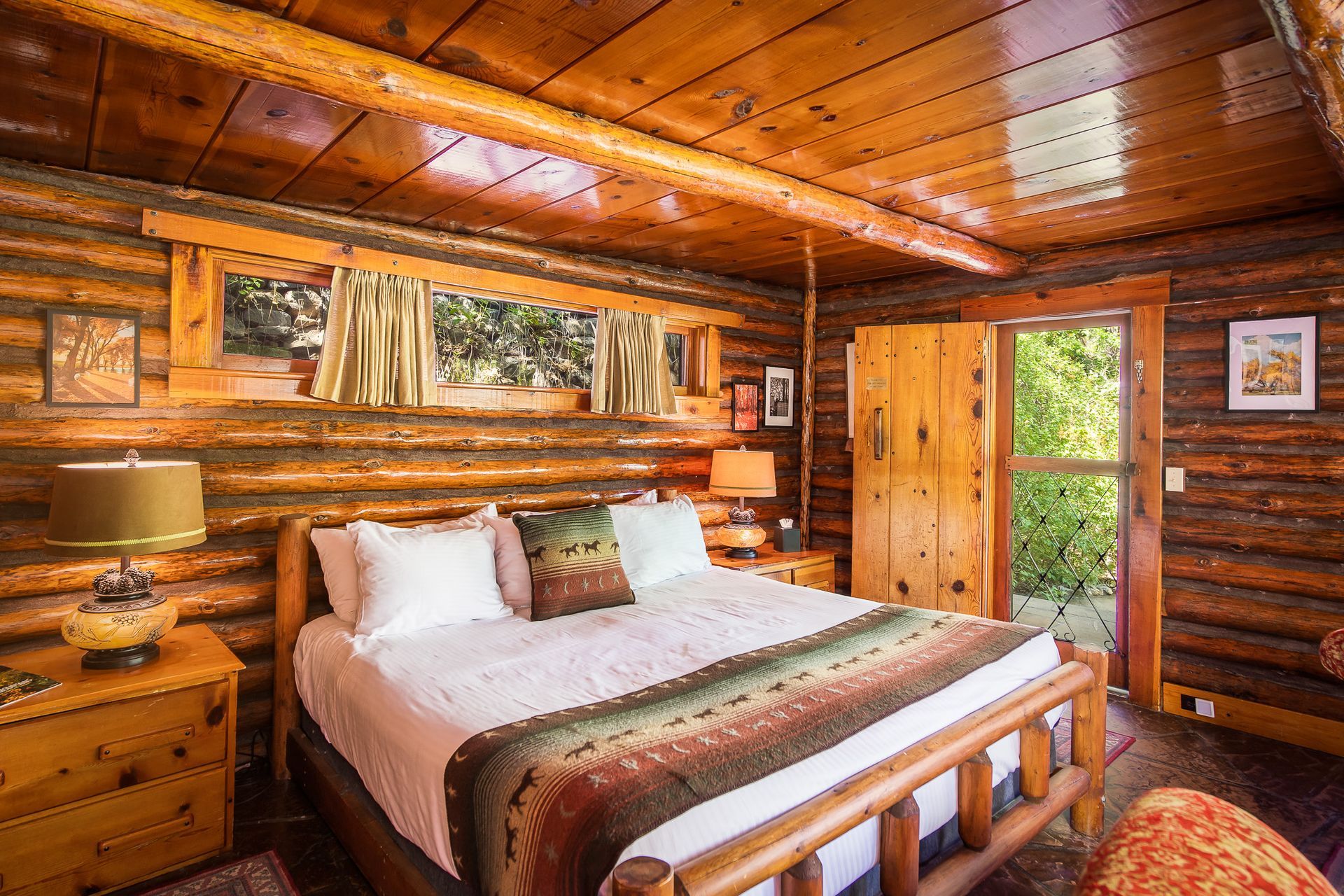 Cozy log cabin bedroom with wooden walls, double bed, bedside lamp, and forest view through the window.