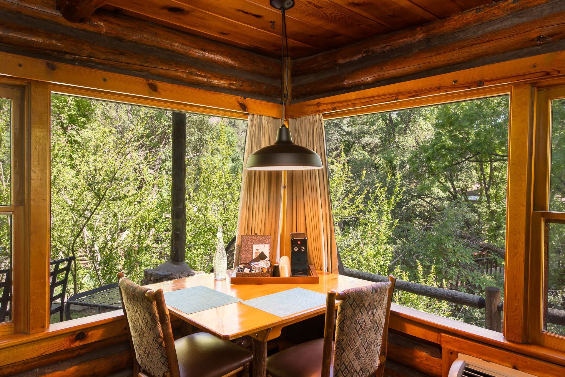 Cozy wooden dining nook with large windows, a table, four chairs, and a hanging pendant light overlooking trees