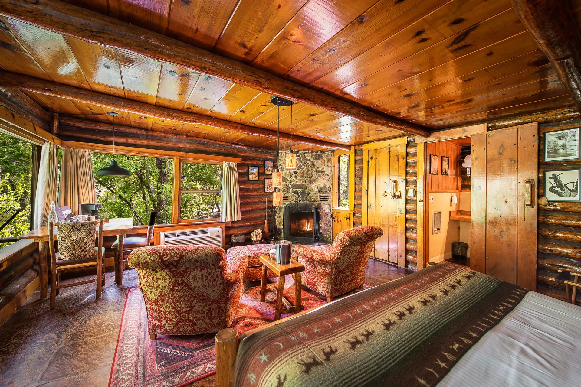 Cozy rustic cabin living room with wood-paneled walls, fireplace, and large windows overlooking greenery