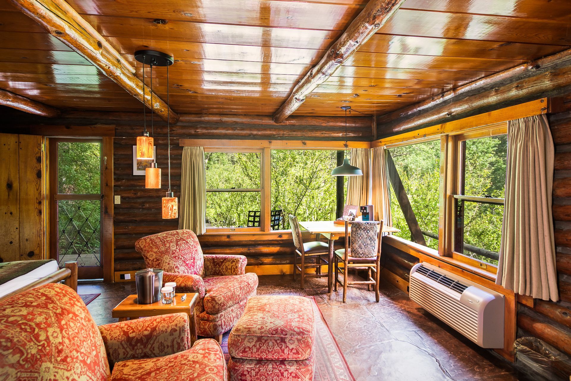 Cozy rustic cabin living room with wood beams, patterned chairs, and large windows overlooking trees