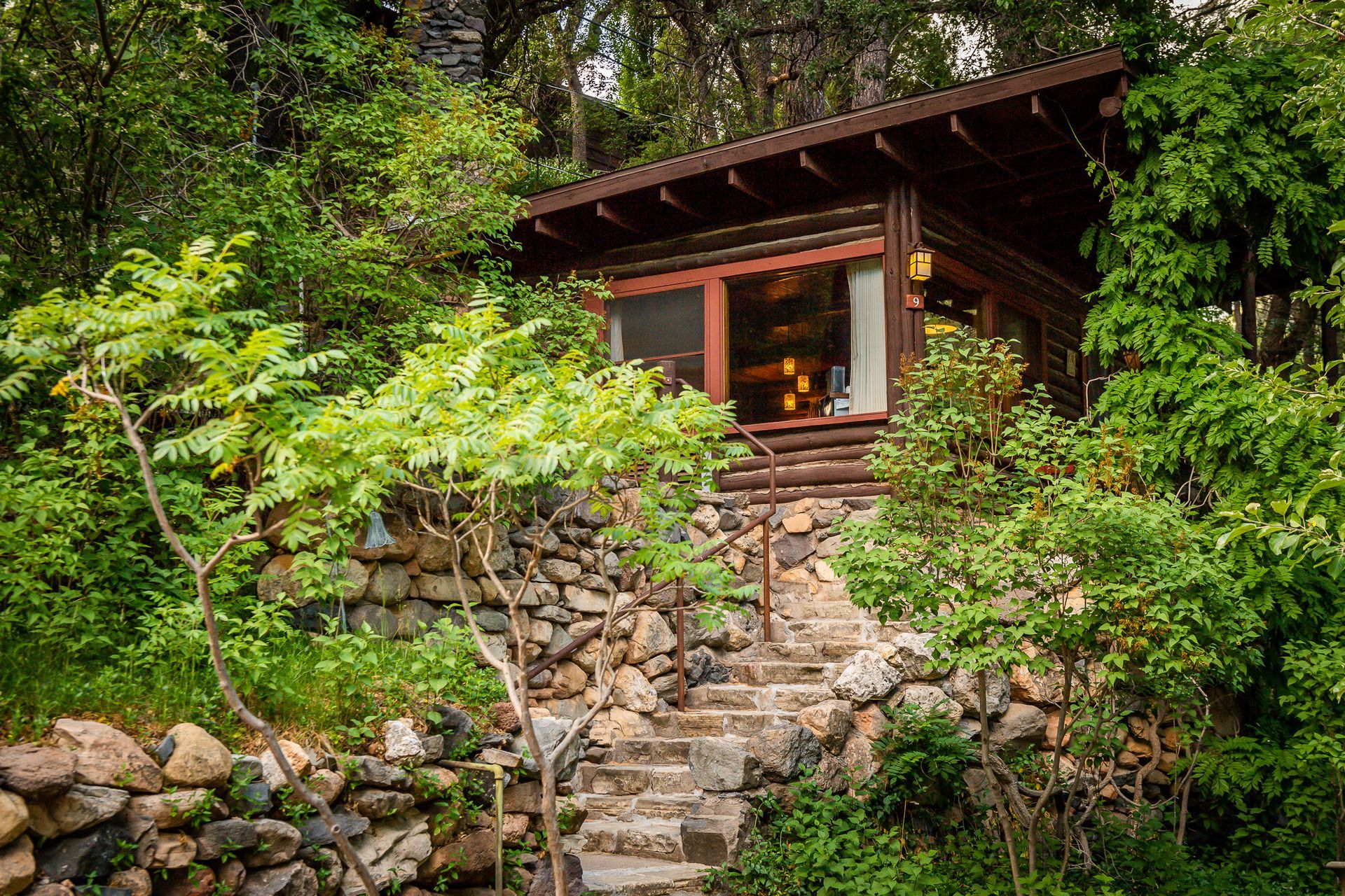 Small rustic cabin on a stone terrace, surrounded by lush green forest and stone steps.