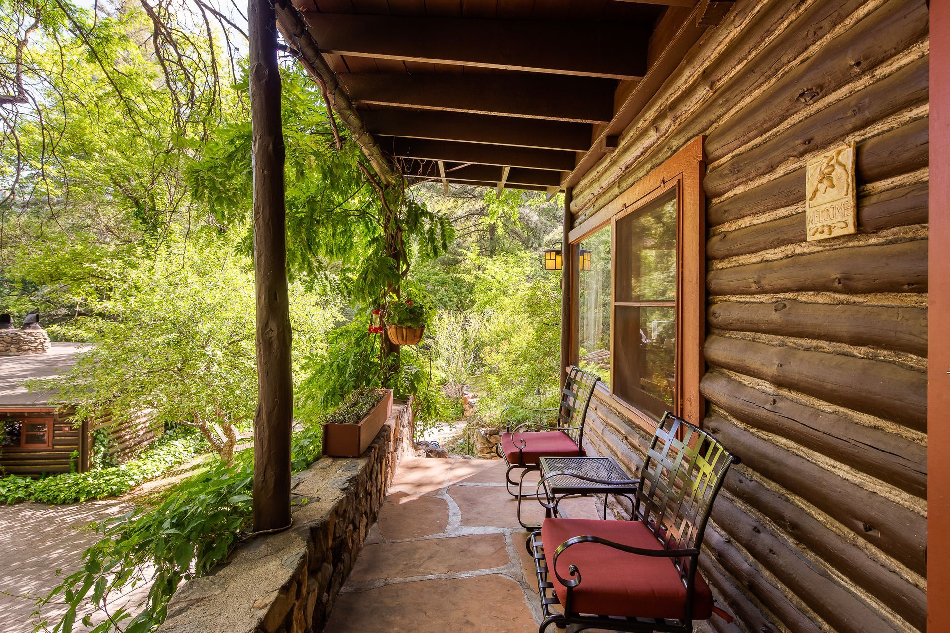Rustic wooden porch with red chairs beside a log cabin, overlooking a leafy yard.