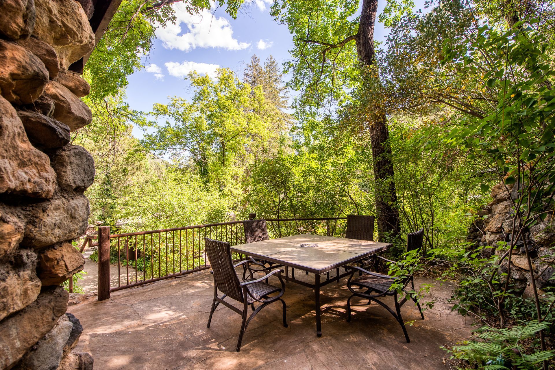 Patio table and chairs on a shaded stone terrace overlooking a bright green wooded landscape
