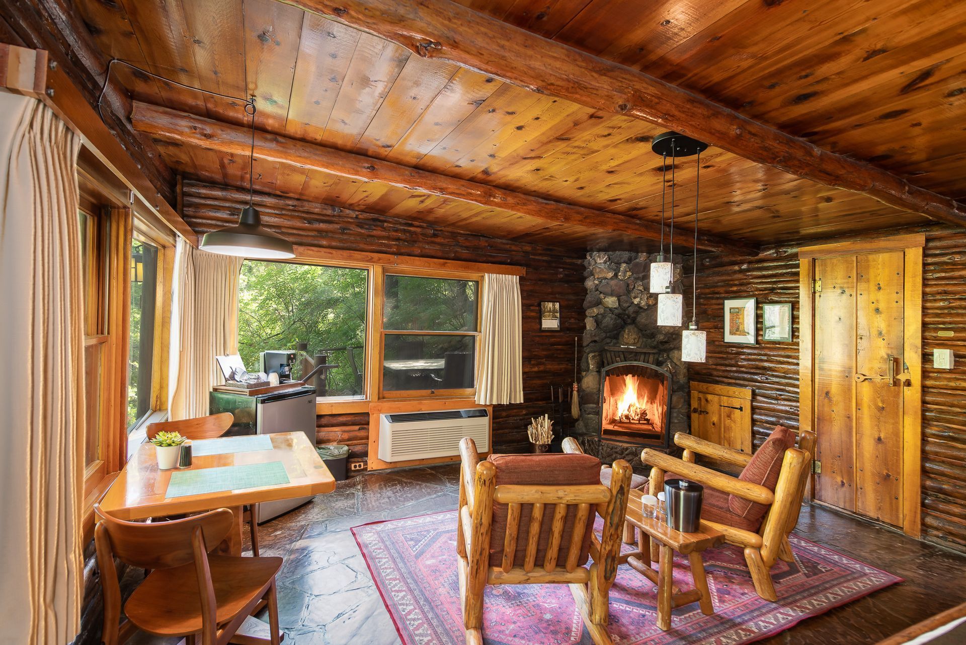 Cozy log cabin living room with wood walls, fireplace, large windows, and rustic chairs on a patterned rug