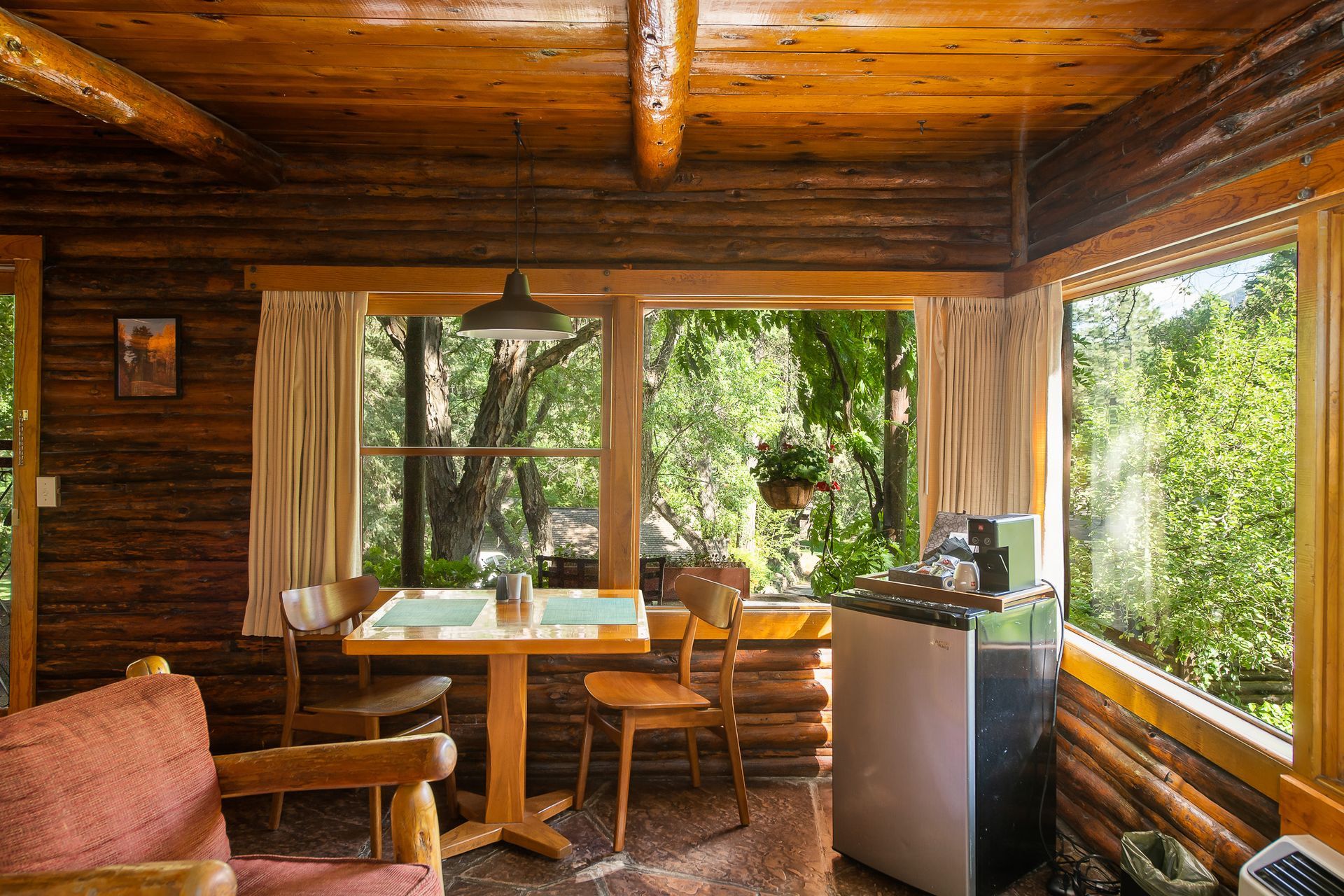 Rustic wood-paneled porch with dining table, chairs, and leafy garden view through open windows
