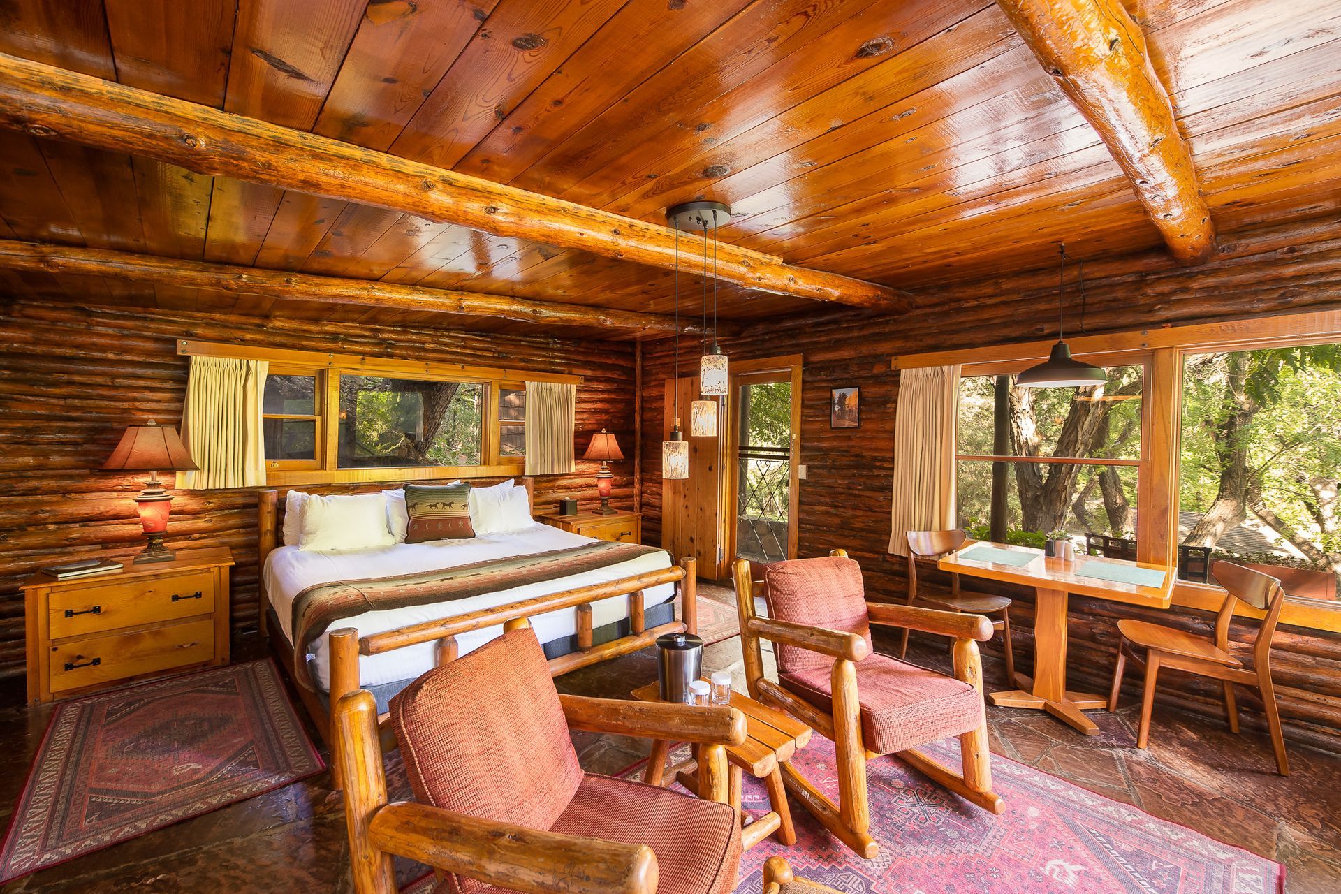 Rustic log cabin bedroom with wooden ceiling, large bed, armchairs, and forest-view windows