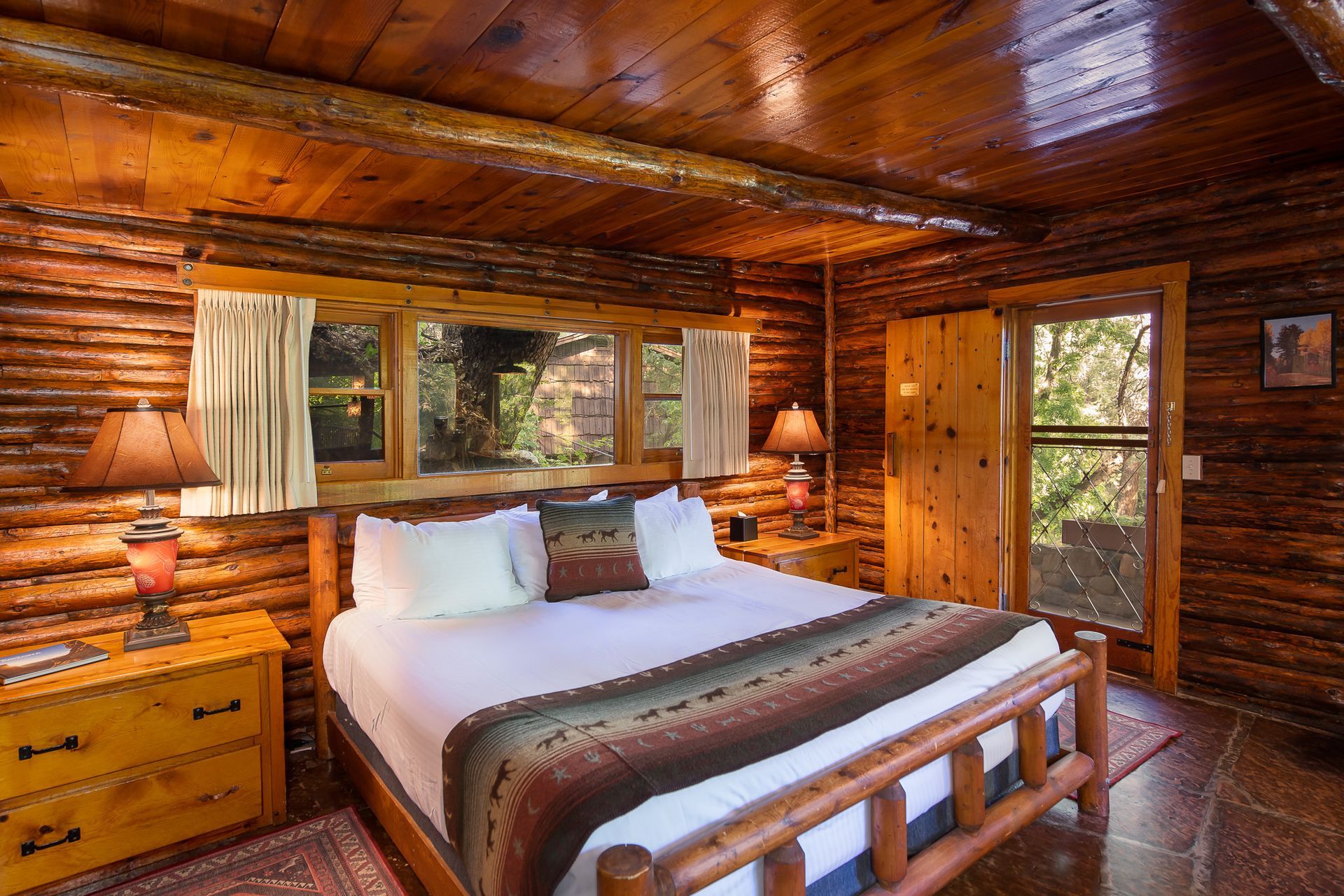Cozy log cabin bedroom with wooden walls, a large bed, lamps, and windows overlooking trees