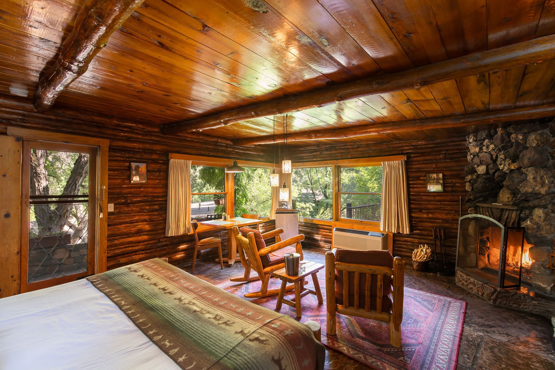 Rustic log cabin bedroom with wood-paneled walls, large windows, bed, chairs, and stone fireplace overlooking forest views