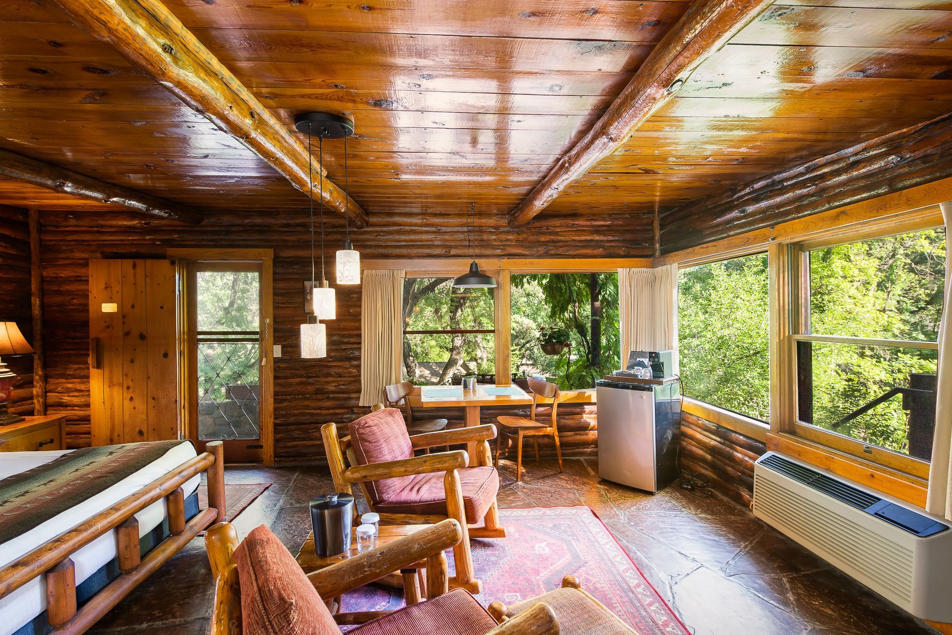 Cozy wood-paneled sunroom with large windows, patterned rug, chairs, and leafy forest view