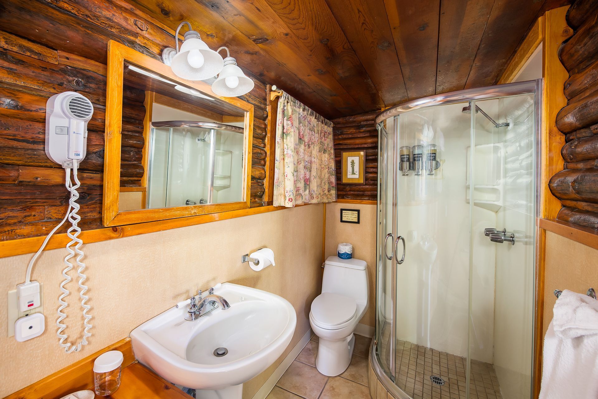 Compact bathroom with wood ceiling, sink, toilet, glass shower, and wall mirror in warm tones