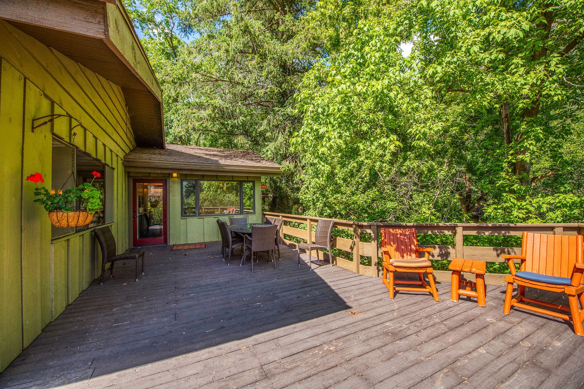 Sunny patio beside yellow house with potted flowers, wooden chairs, and trees in the background