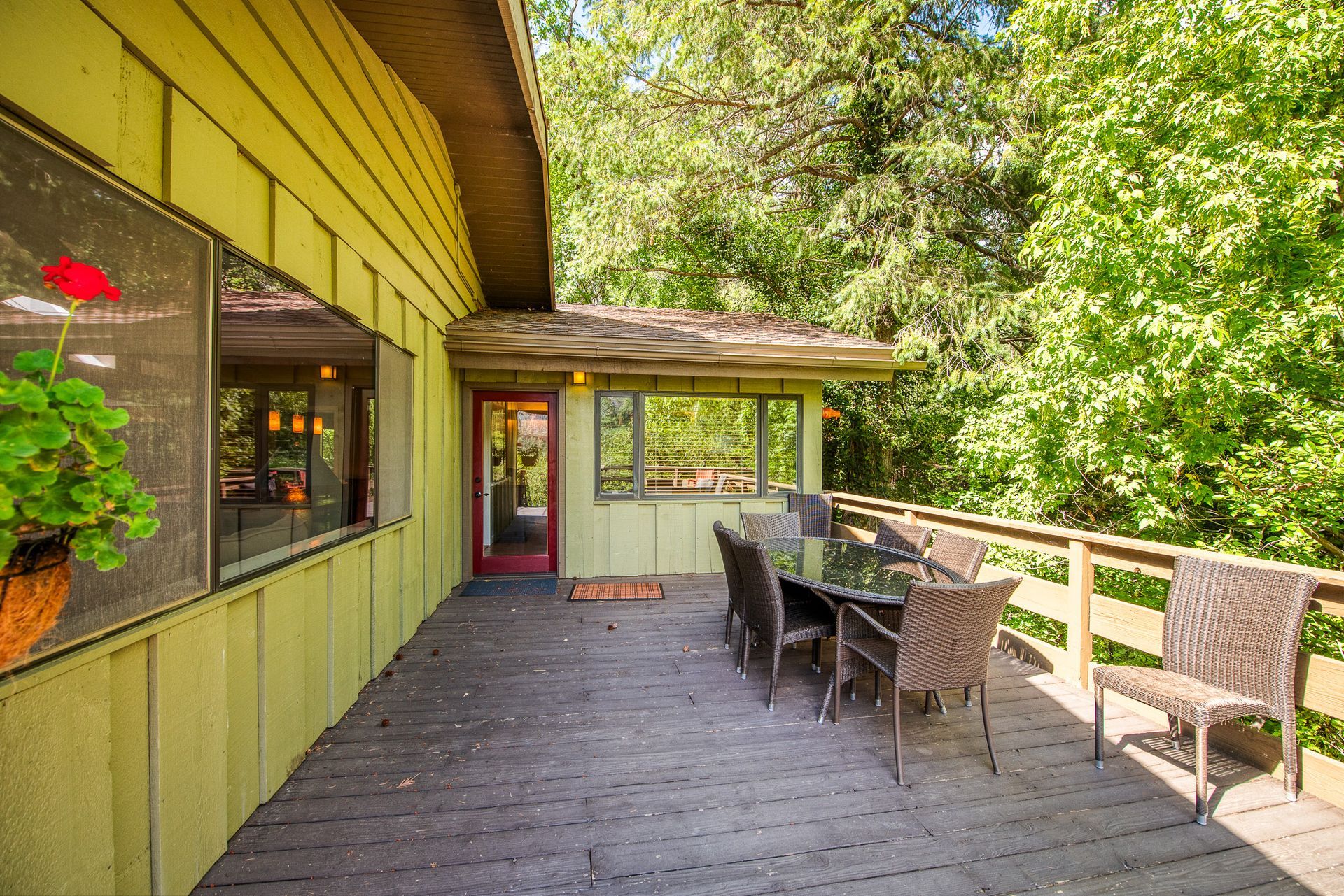 Covered wooden patio with table, chairs, potted plants, and green siding beside a wooded view.