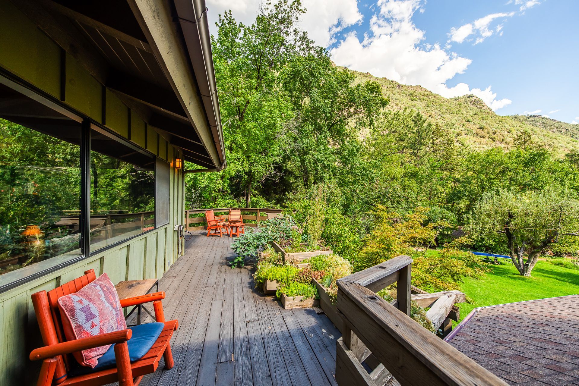 Wooden deck with orange chairs beside a house, overlooking lush green trees and a sunny hillside.