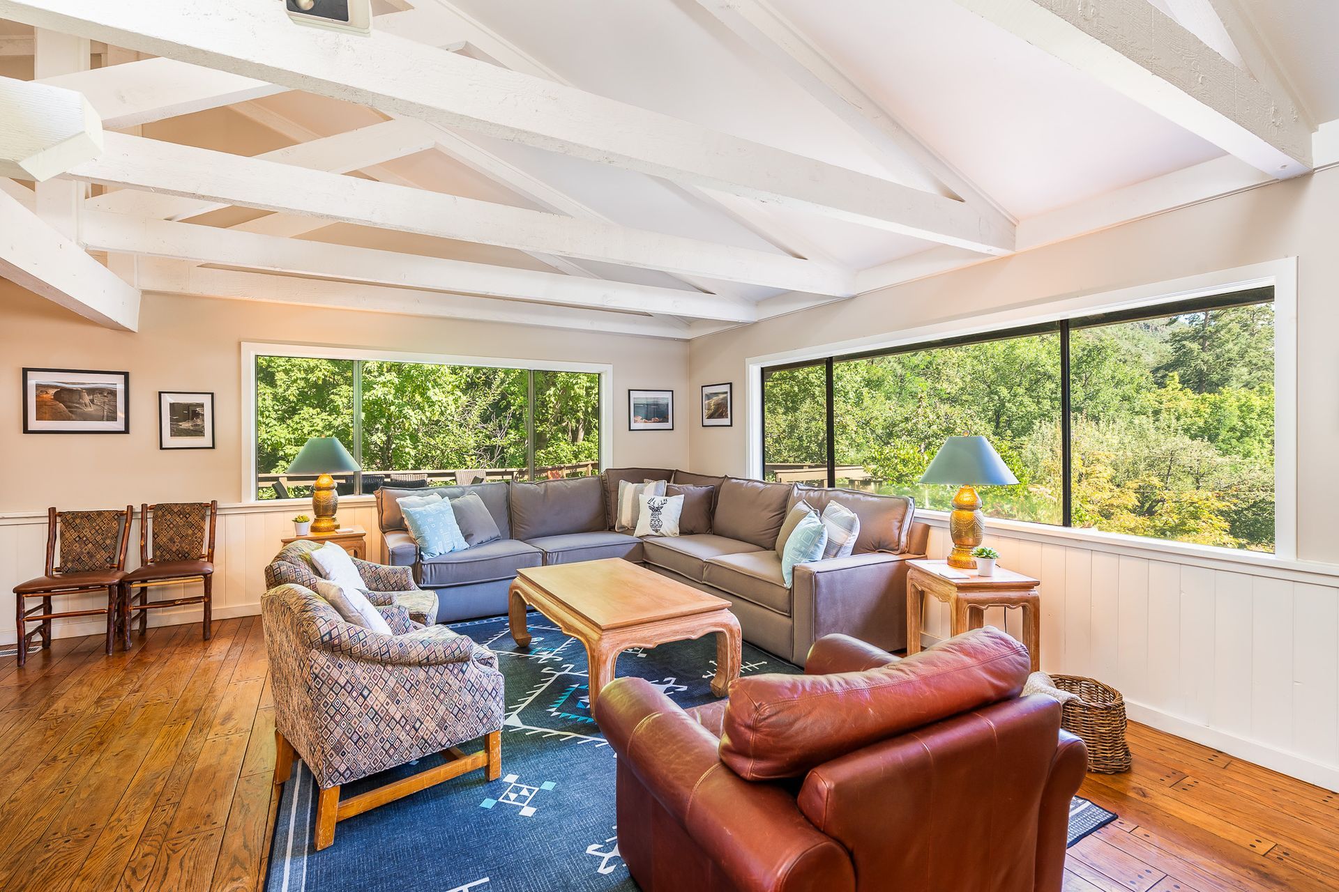 Bright living room with blue sofa, leather chair, wooden table, and large windows overlooking greenery