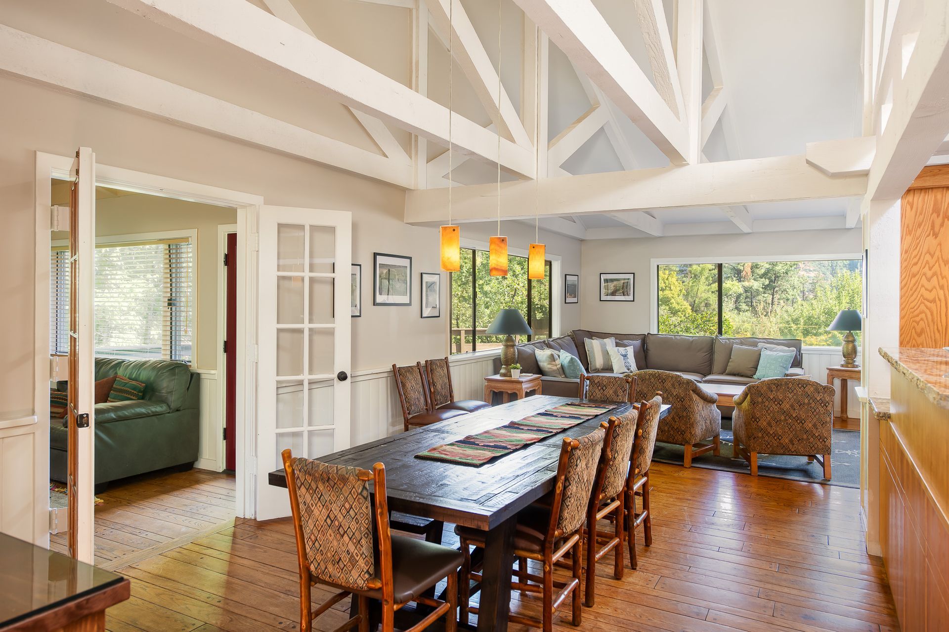 Bright dining room with exposed beams, long wooden table, wicker chairs, and large windows overlooking greenery