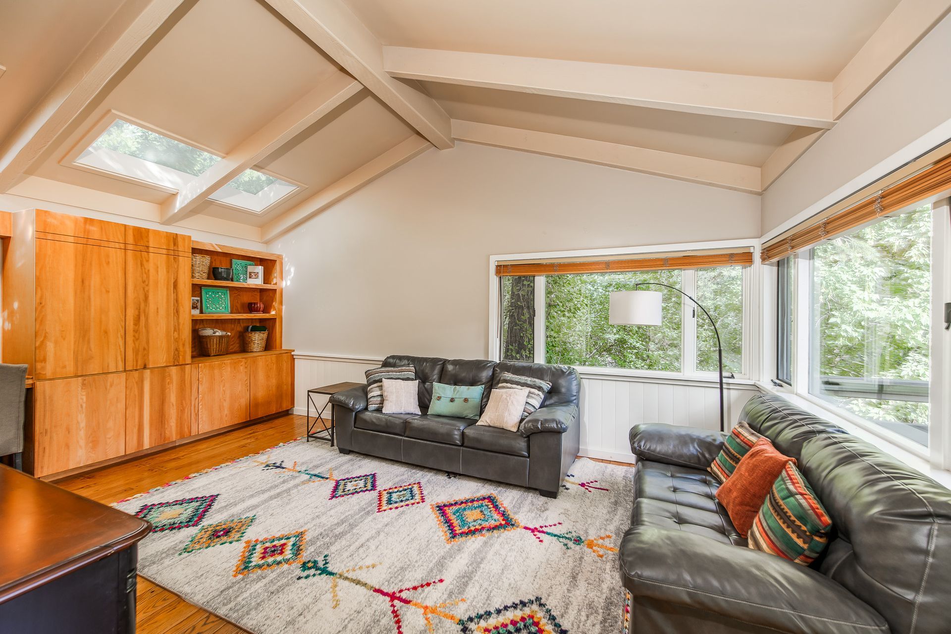 Bright living room with vaulted ceiling, large windows, leather sofa, patterned rug, and wooden cabinet