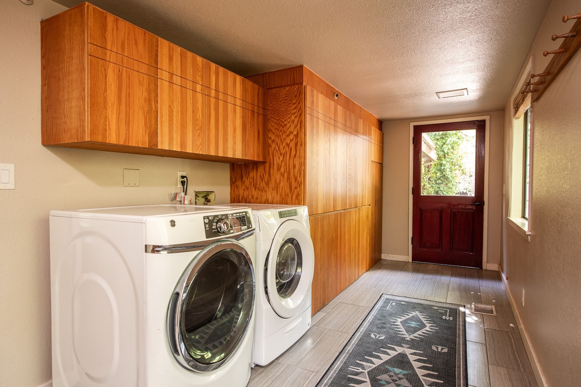 Laundry room with white washer and dryer, wood cabinets, and a patterned runner leading to a red door.